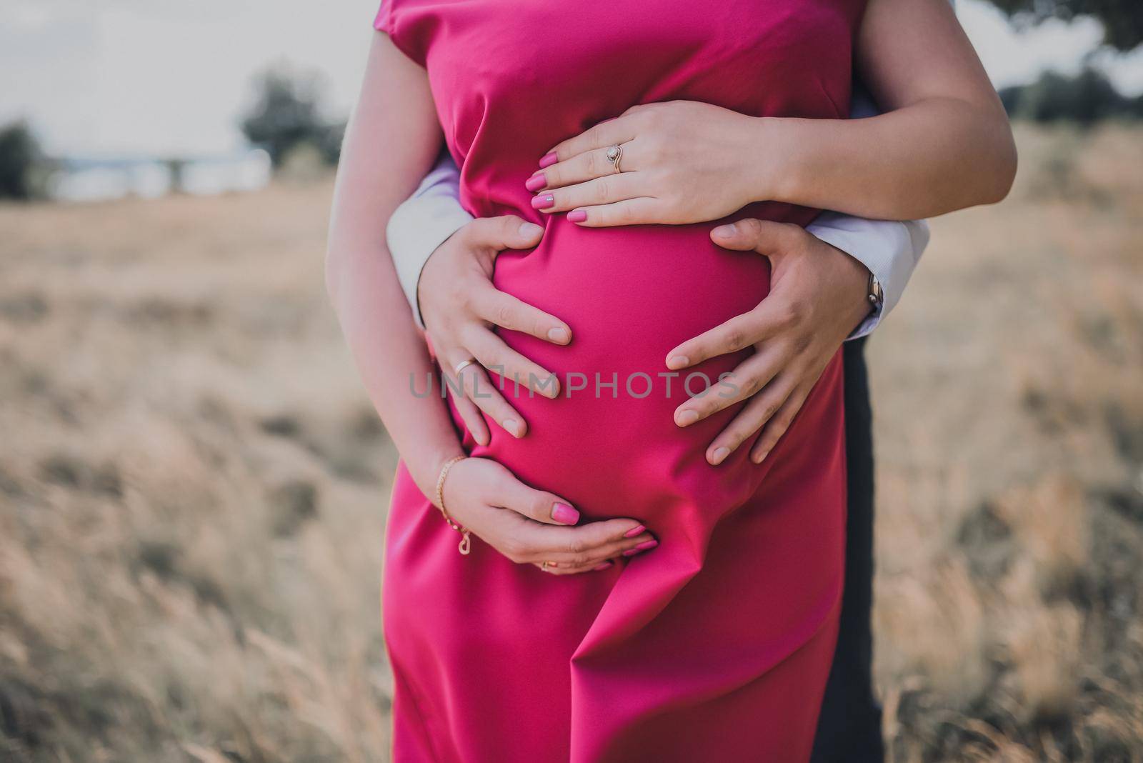 Male and female hands hug a pregnant belly. Pregnant european girl in a pink dress. against the background of summer nature and the river