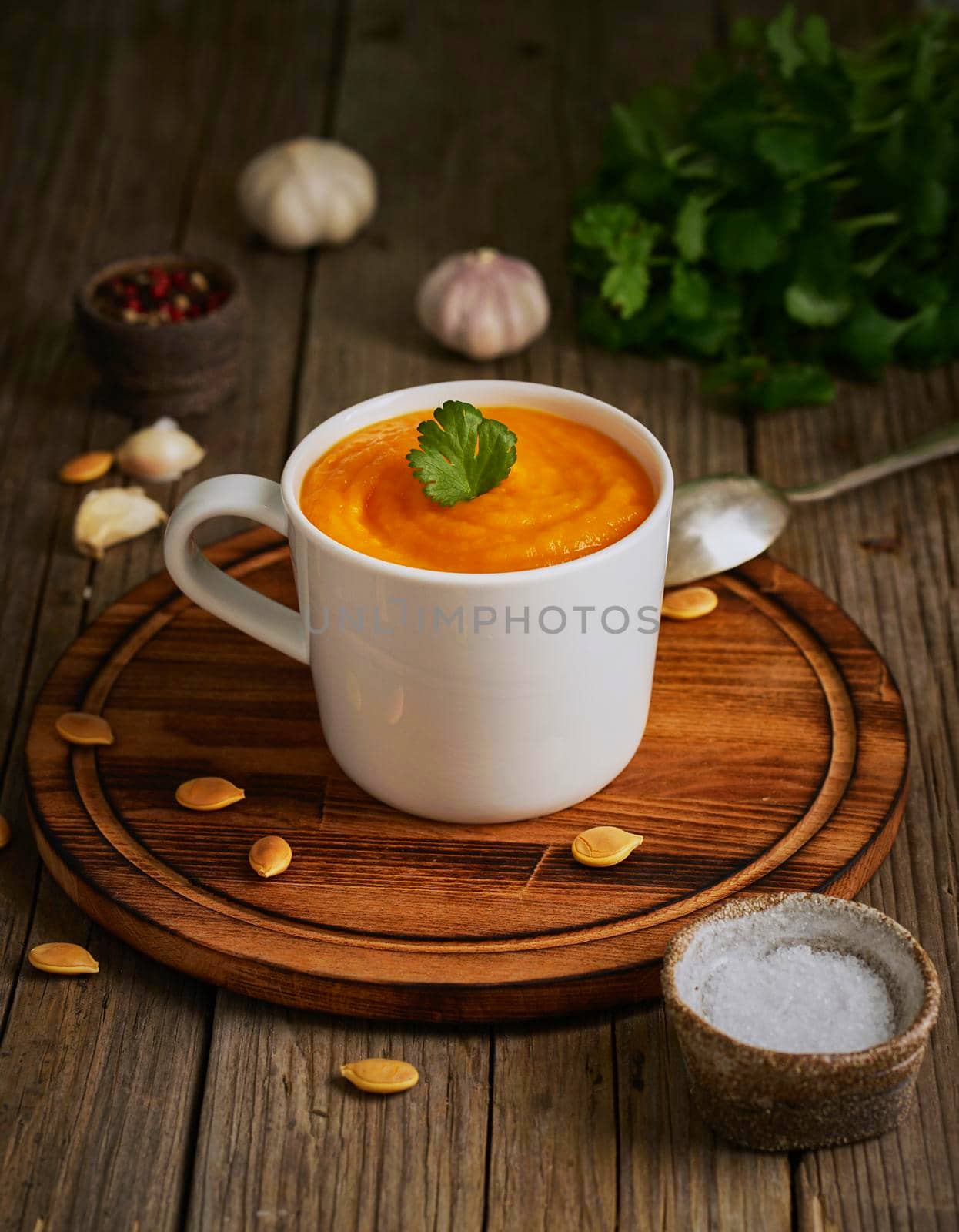 Pupmkin cream soup in cup on brown wooden table, vertical, side view. Dietary vegetarian puree on cutting board with parsley, garlic.