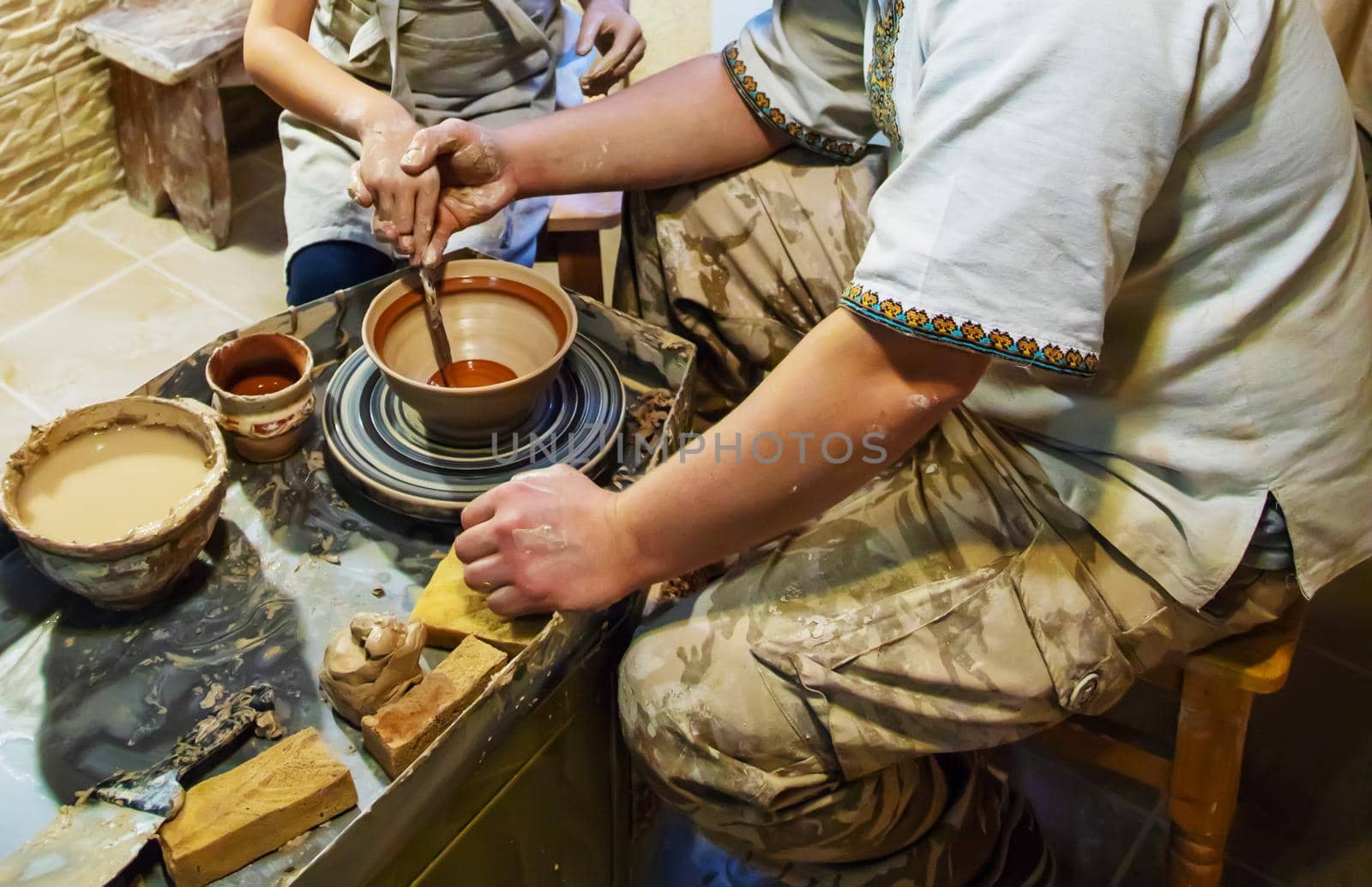 the Hands of a master and a student make a pitcher on a Potters wheel of yellow clay. Selective focus on hands