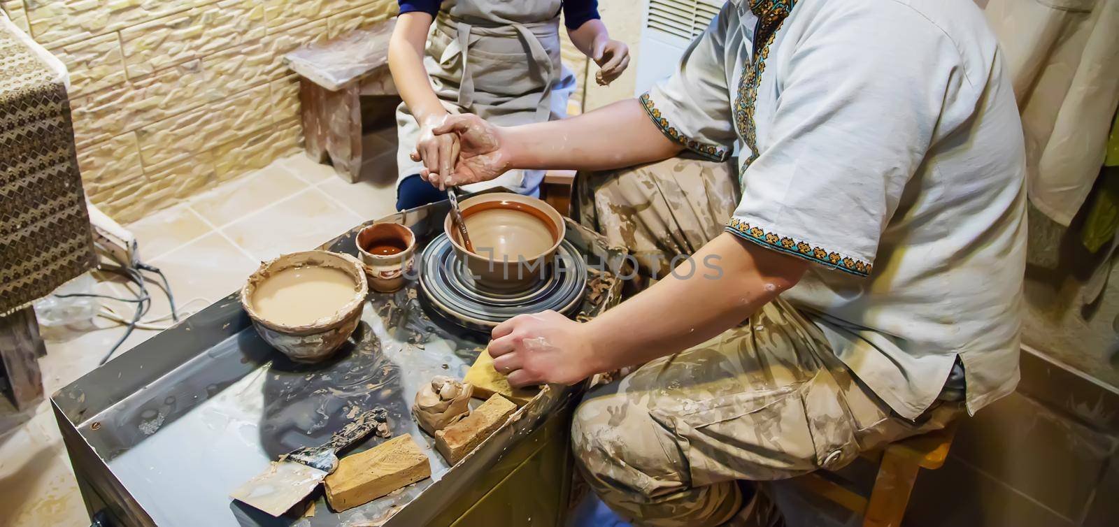 the Hands of a master and a student make a pitcher on a Potters wheel of yellow clay. Selective focus on hands by mila1784