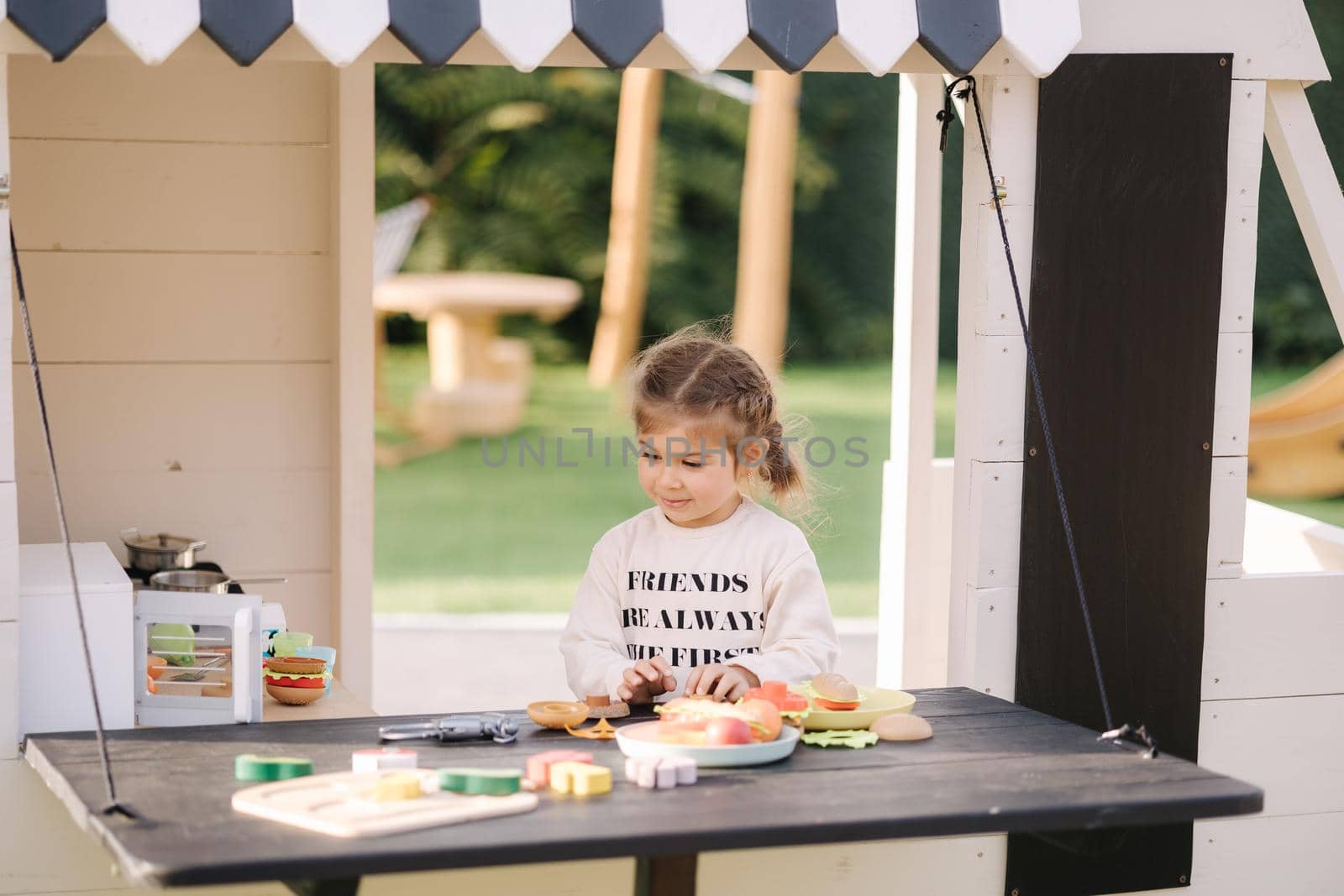 Happy little girl playing on toy kitchen on wheels. Cute girl make a burger on toy kitchen outdoors. Adorable kid play on fresh air.