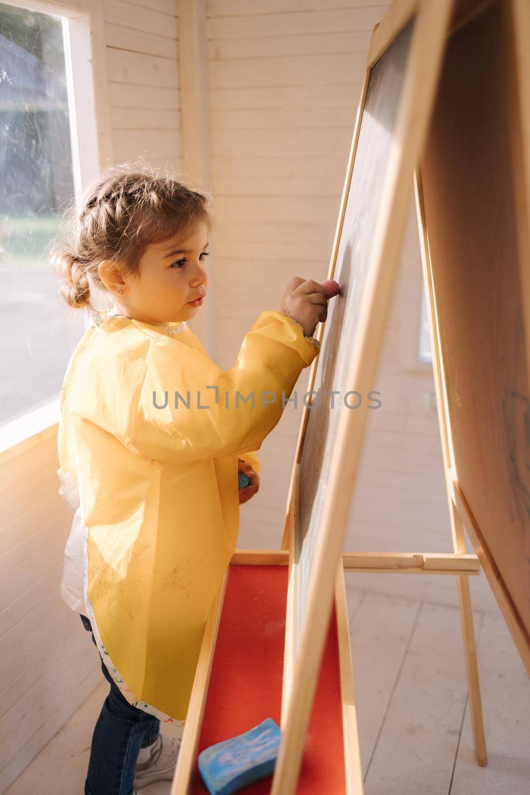 Young artist paints on wooden boards with chalk. Cute girl in yeallow raincoat.