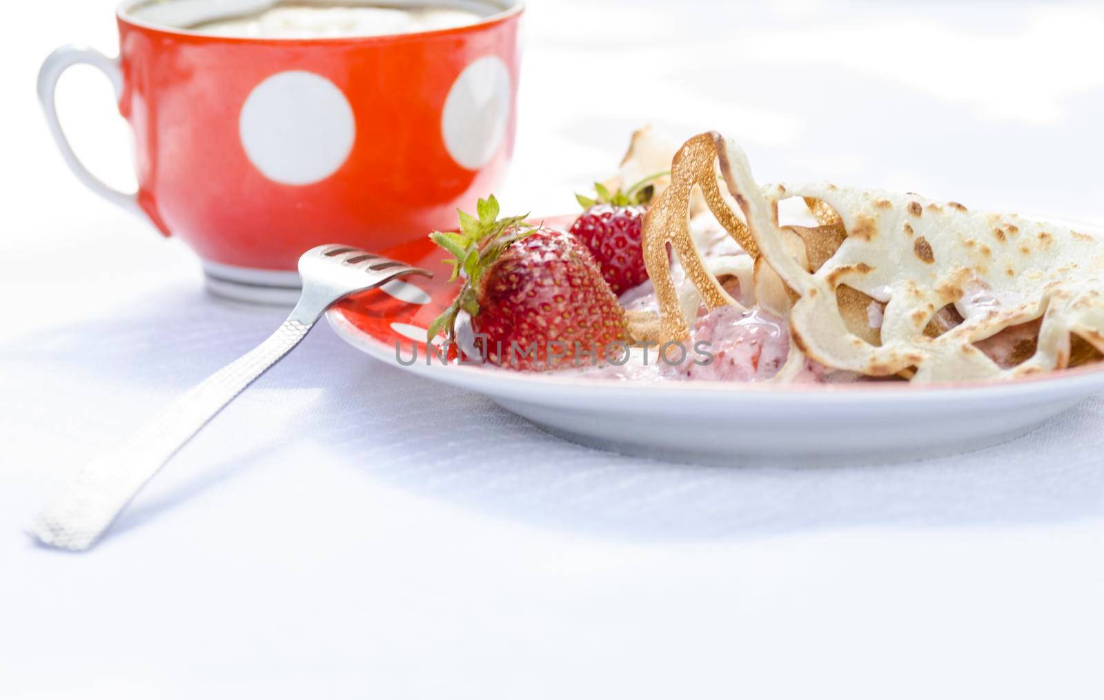 Pancakes with strawberries and sour cream on the white tablecloth. Next to a cup of coffee. All lit bright sun. On the tablecloth visible shadows from tree leaves.