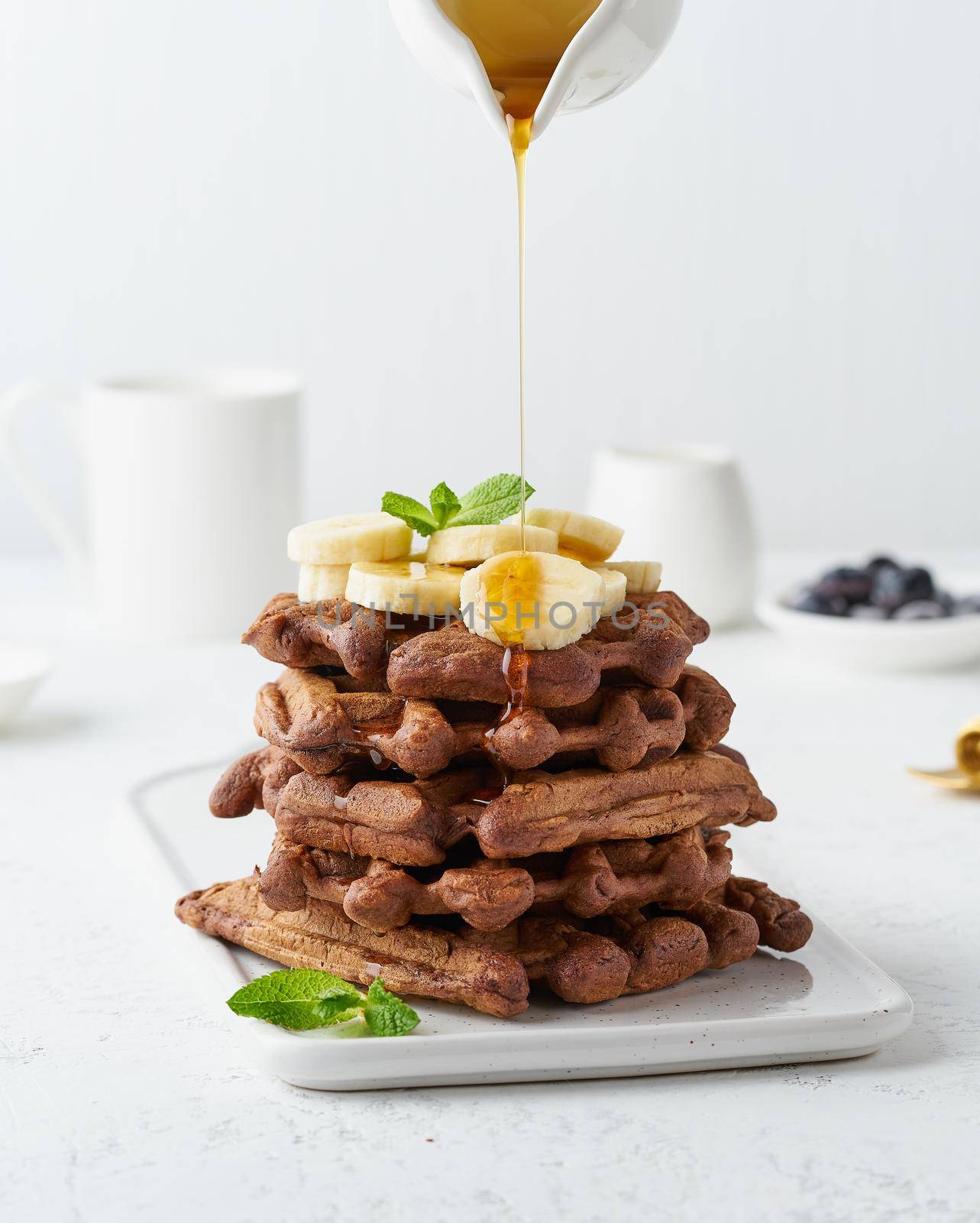 Chocolate banana waffles with maple syrup on white table, side view, vertical. Sweet brunch, maple syrup flow in milk jug, a creamer.