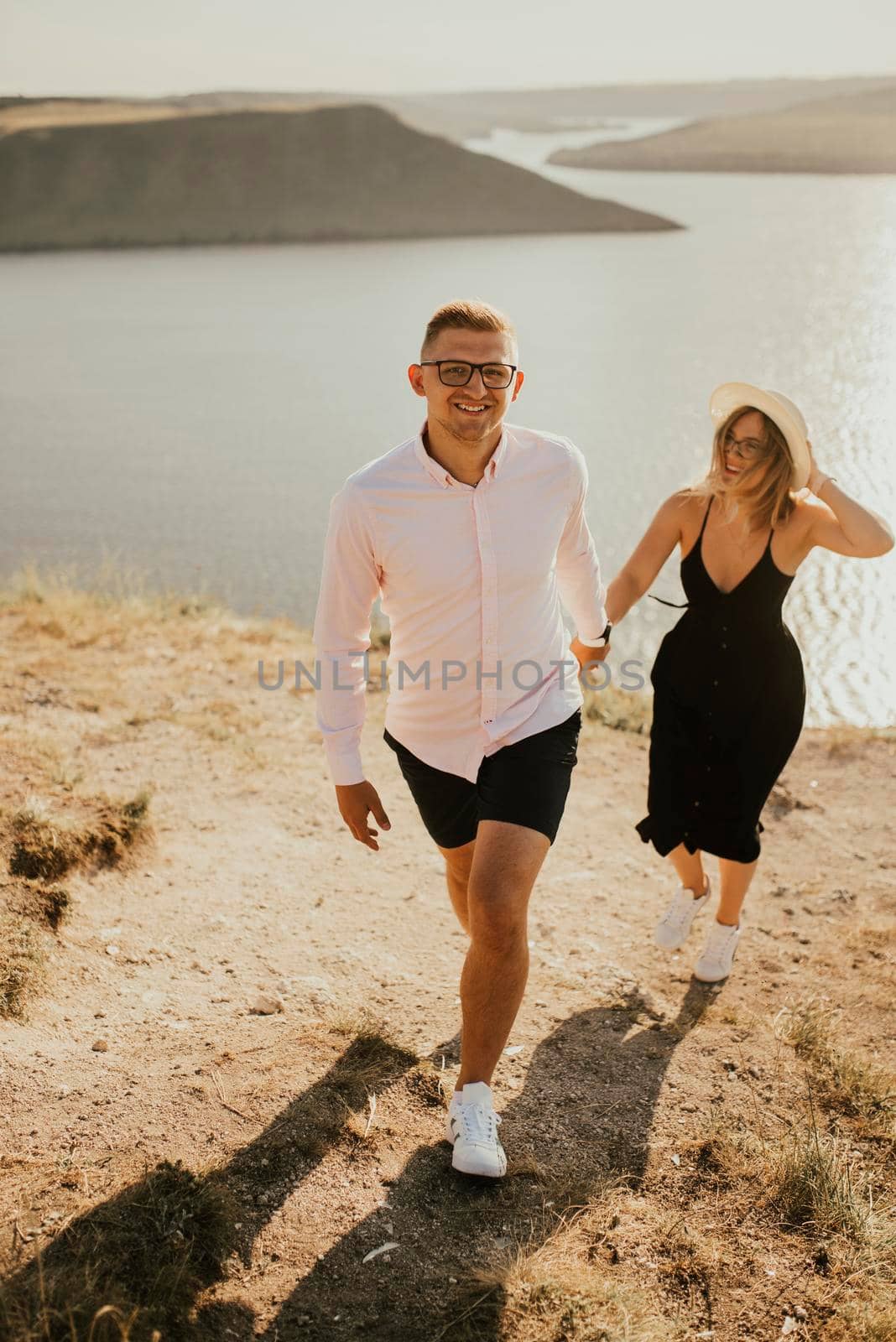 A man and a woman in love walk on a mountain above a large lake by the sea at sunset. A couple of fair-haired fair-skinned people in love are resting in nature in a field