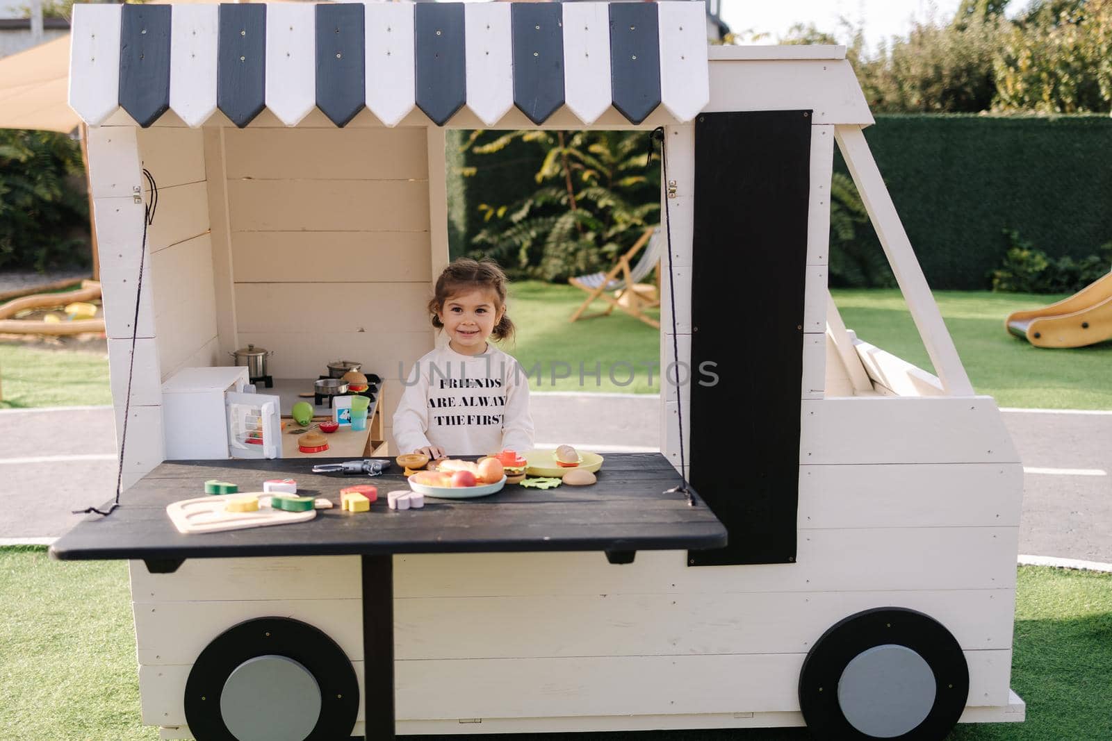 Happy little girl playing on toy kitchen on wheels. Cute girl make a burger on toy kitchen outdoors. Adorable kid play on fresh air.