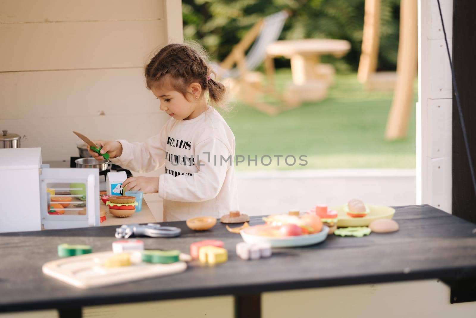 Happy little girl playing on toy kitchen on wheels. Cute girl make a burger on toy kitchen outdoors. Adorable kid play on fresh air.