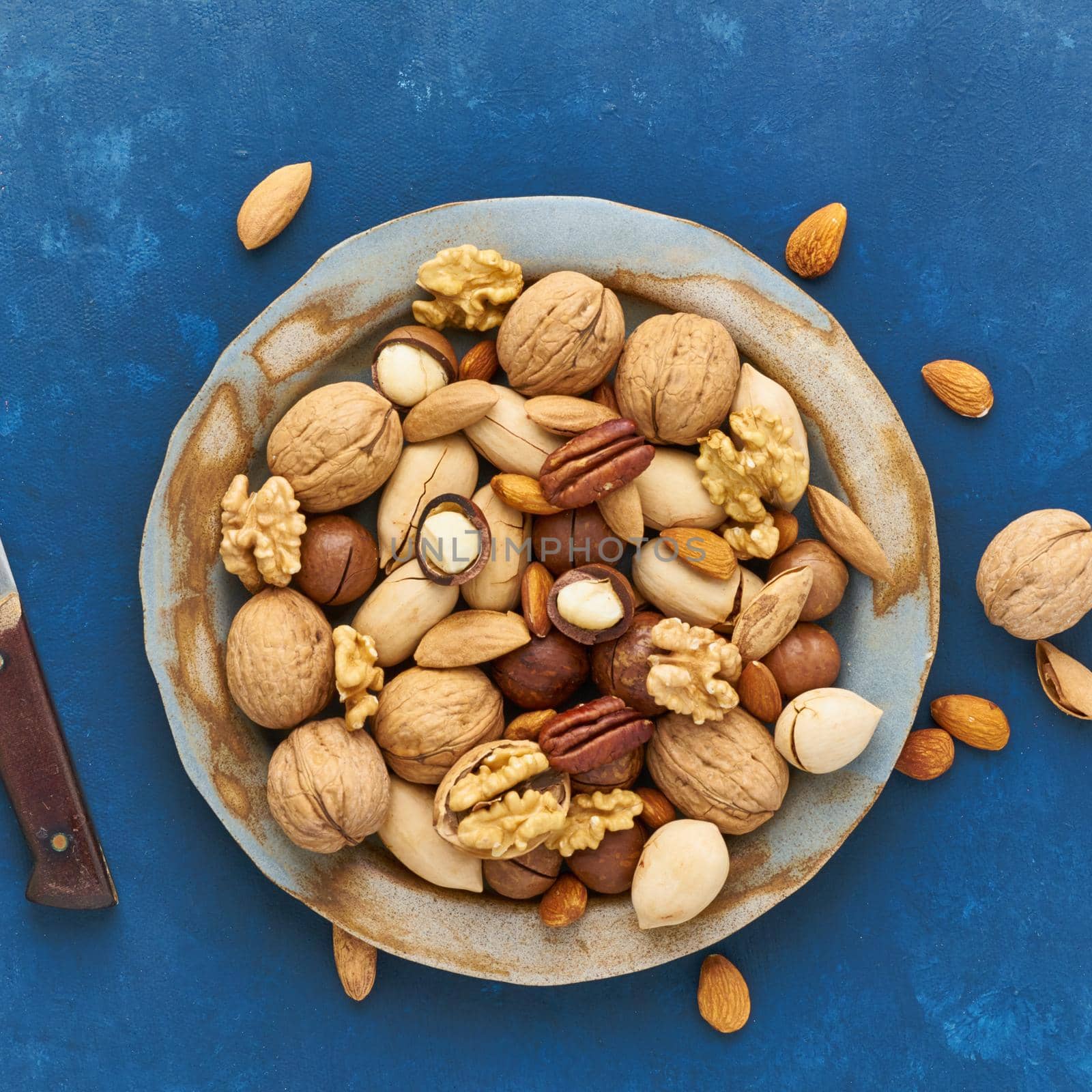 Classic blue in a food. Mix of nuts on plate - walnut, almonds, pecans, macadamia and knife for opening shell. Healthy vegan food. Clean eating, balanced diet. Top view, close up