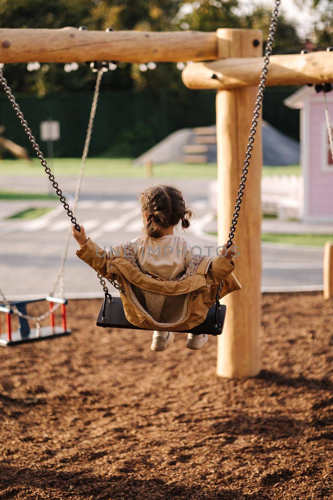 Happy little girl swing on a swing in the specially designated place in the park. Softner from sawdust underfoot.