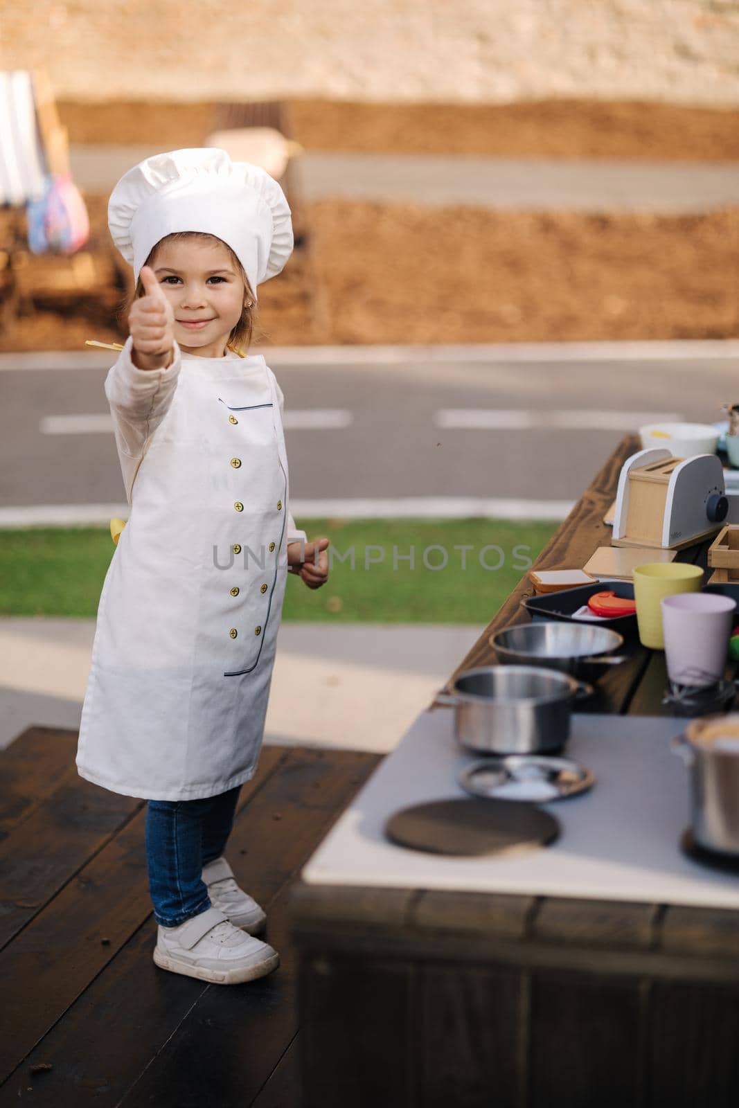 Adorable little girl in chef's coat and cap cooks at the children's toy kitchen. Playing on little kids city.