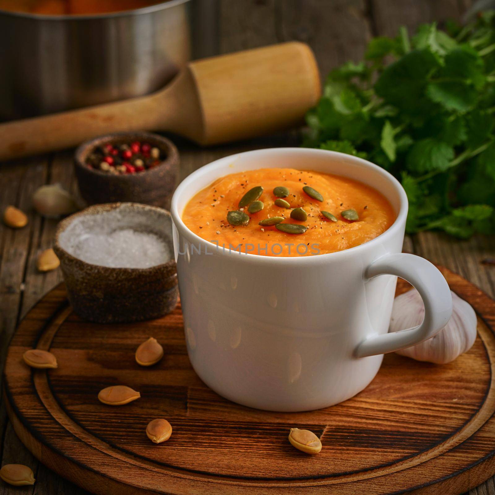 Pupmkin cream soup in cup on brown wooden table, side view. Dietary vegetarian puree on cutting board with parsley, garlic.