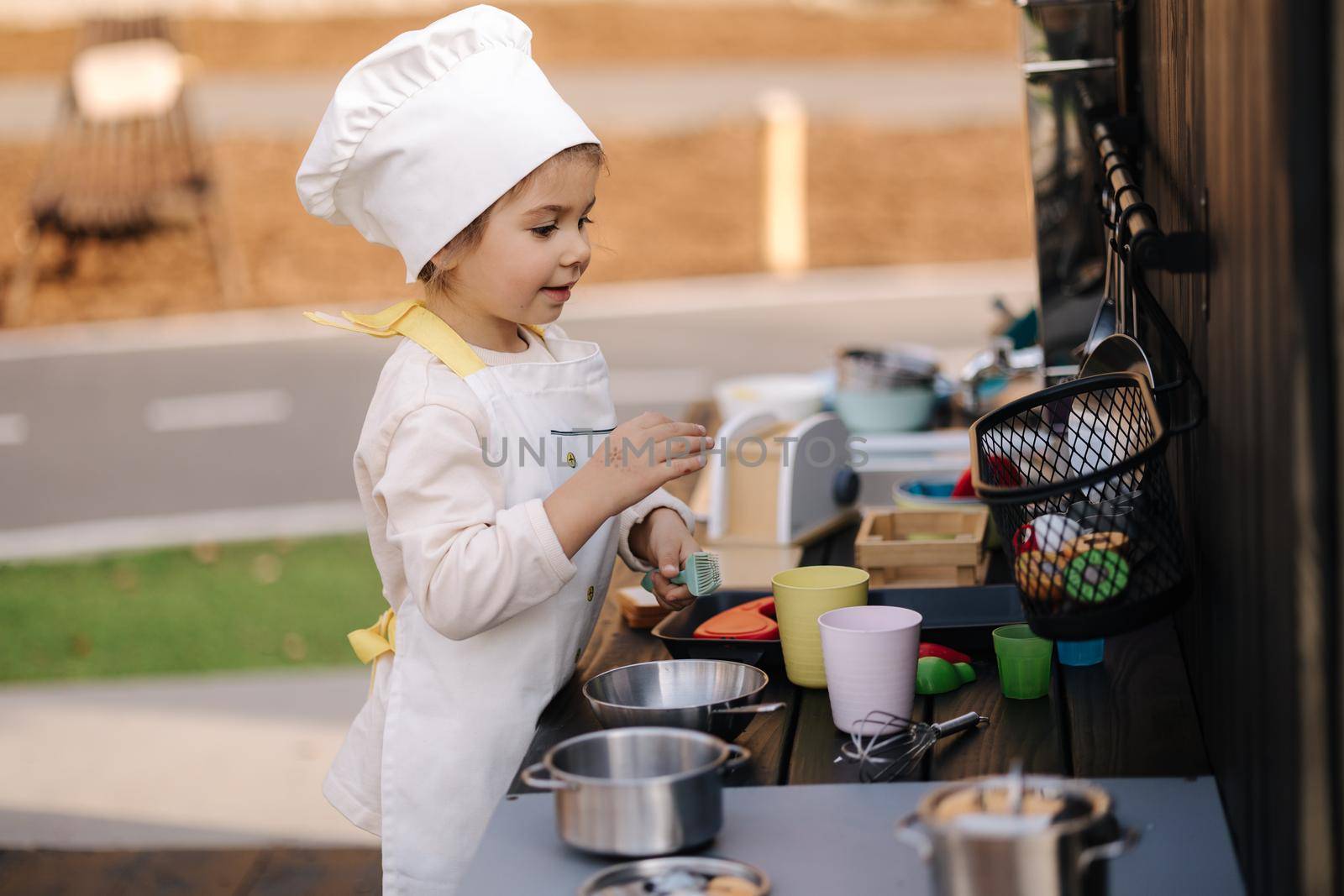 Adorable little girl in chef's coat and cap cooks at the children's toy kitchen. Playing on little kids city.