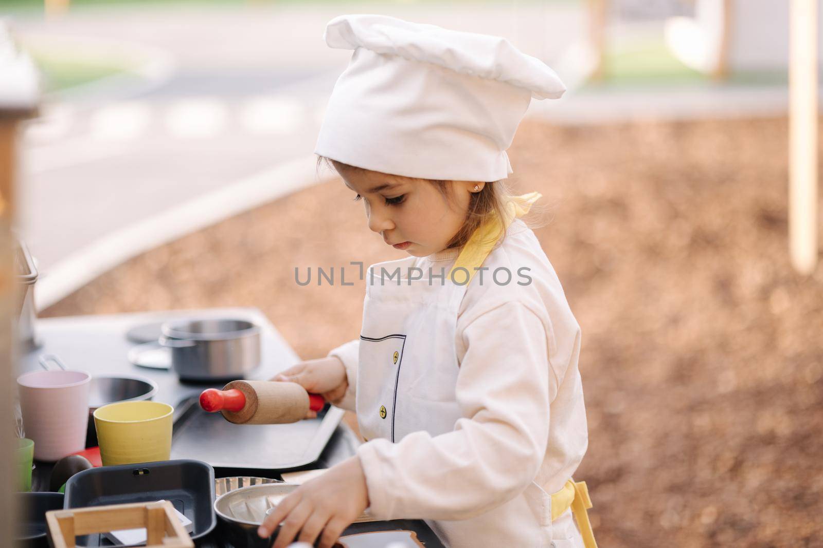 Adorable little girl in chef's coat and cap cooks at the children's toy kitchen. Playing on little kids city.