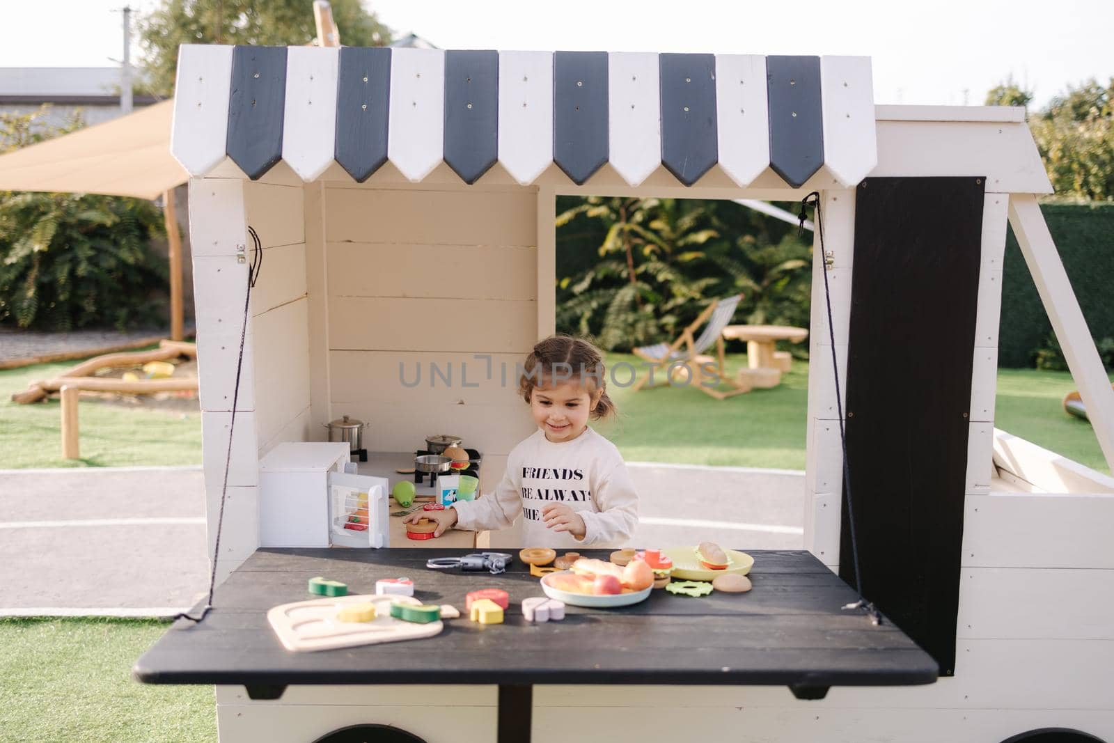 Happy little girl playing on toy kitchen on wheels. Cute girl make a burger on toy kitchen outdoors. Adorable kid play on fresh air.