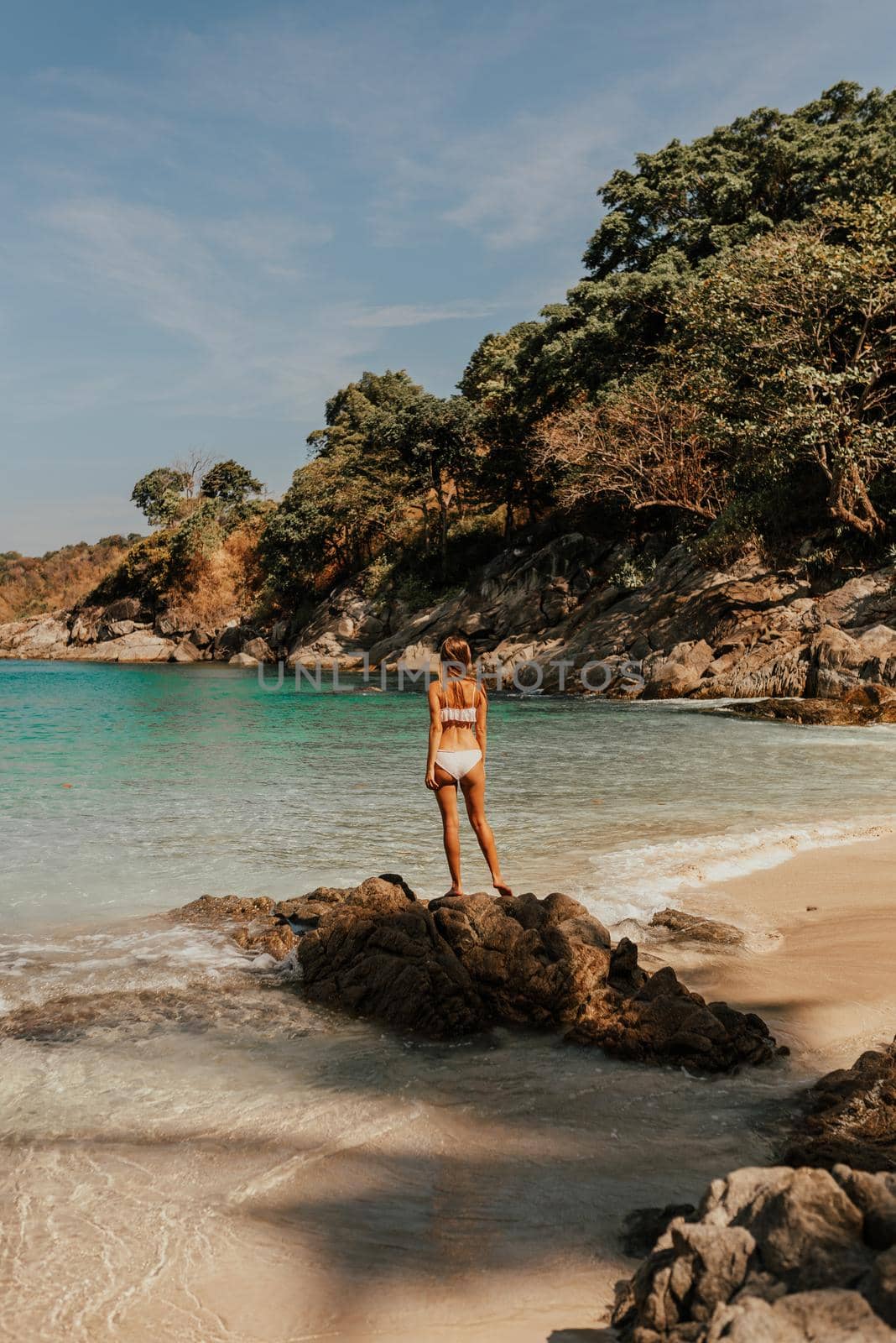 a young blonde European woman in a white bikini swimsuit on the beach stands backwards on the stone rocks on the ocean shore.