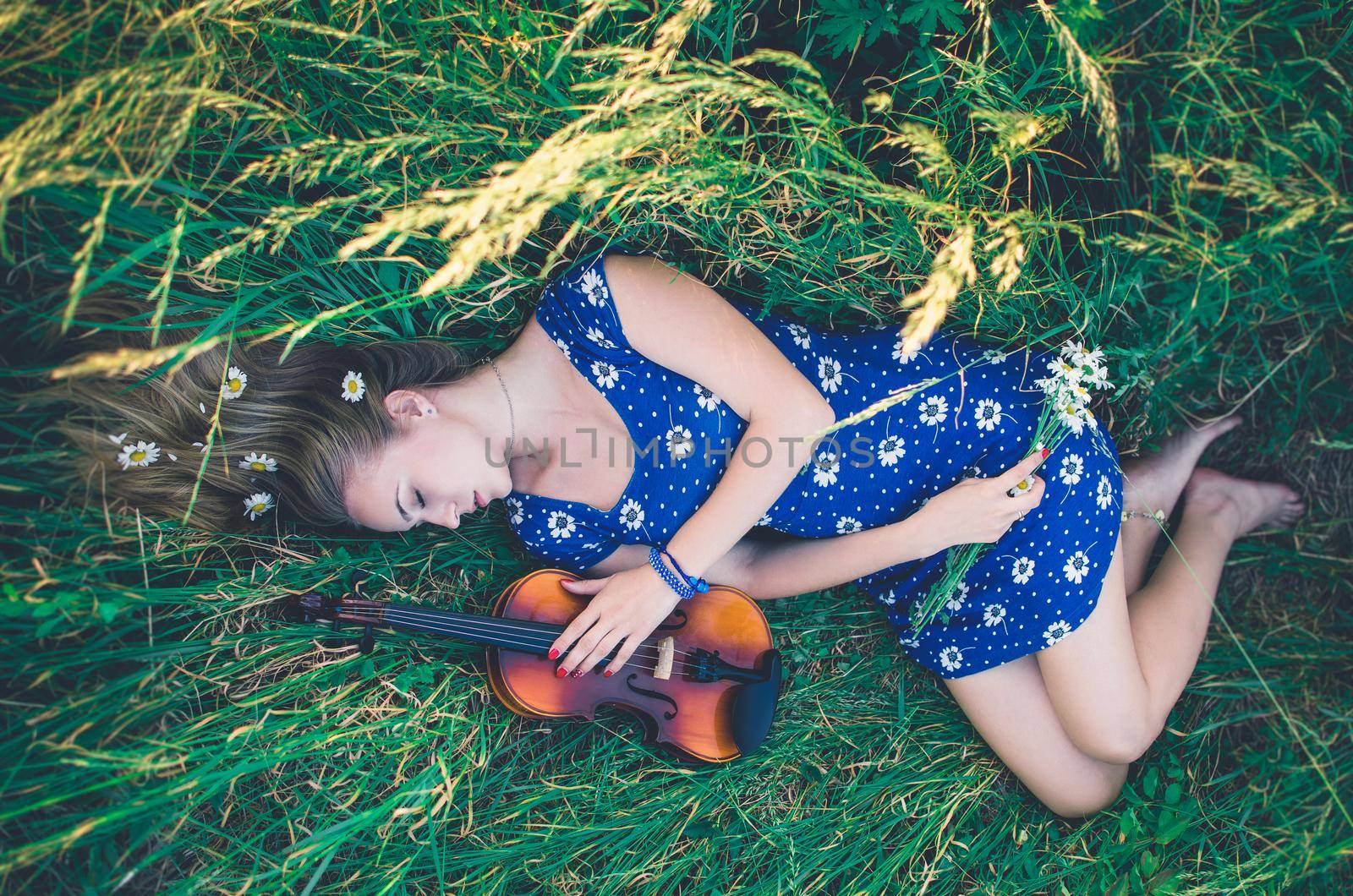 Young slim fair-skinned girl blonde beautiful lies in tall grass. Top view. Girl in a blue short tight dress with a print of daisies. A bouquet and lonely daisies scattered around.A woman hugs violin.