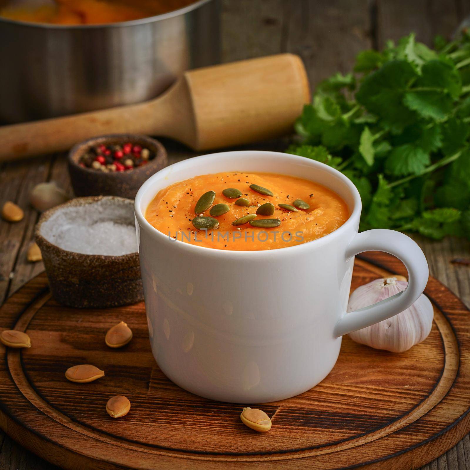 Pupmkin cream soup in cup on brown wooden table, side view. Dietary vegetarian puree on cutting board with parsley, garlic.