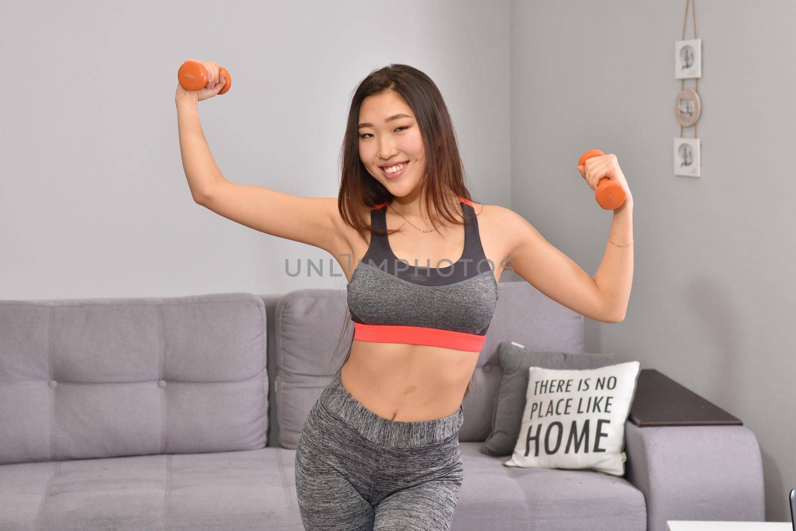 Young asian brunette woman in sportwear making home workout with two orange dumbbells. Soft focus
