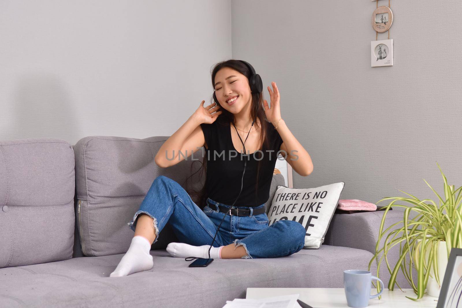 Excited beautiful asian teen listening music in headphones on her sofa at home. Wearing jeans and black tshirt