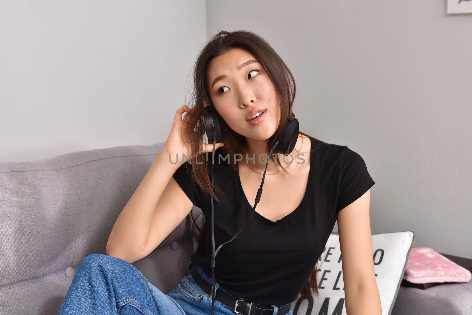 Excited beautiful asian teen listening music in headphones on her sofa at home. Wearing jeans and black tshirt