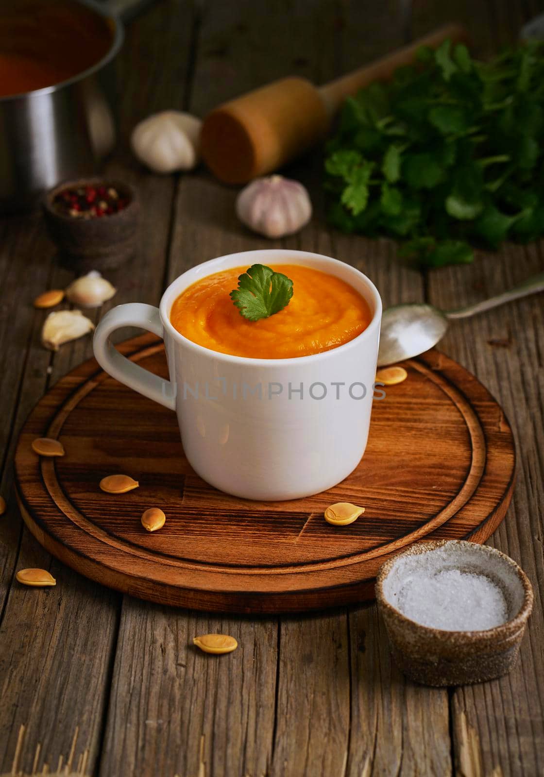 Pupmkin cream soup in cup on brown wooden table, vertical, side view. Dietary vegetarian puree on cutting board with parsley, garlic.