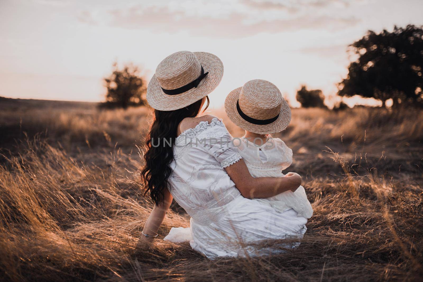 brunette mother and little daughter are sitting on the dry grass in white dresses. On the Sunset. in hats boater with his back.