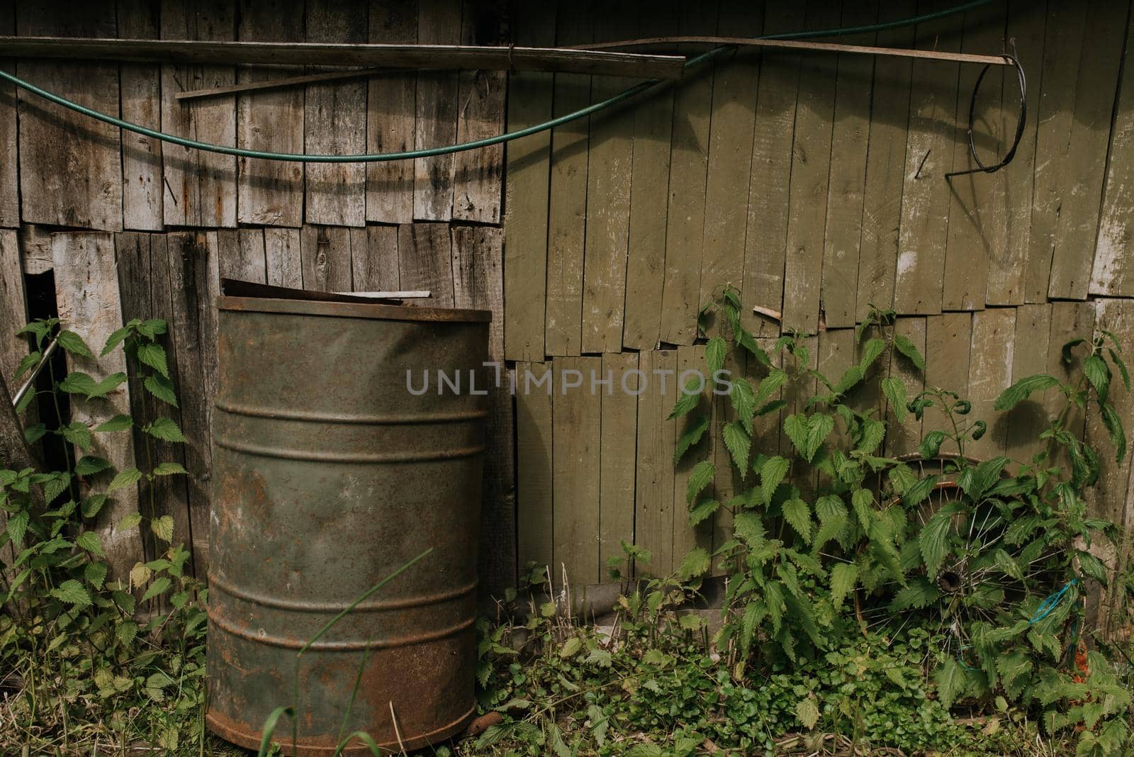 Old wooden shabby frail little fishing house. Under the roof of the house is attached a fishing rod for fishing. Near the house there is a huge Green barrel for water. Old iron metal around. Green big