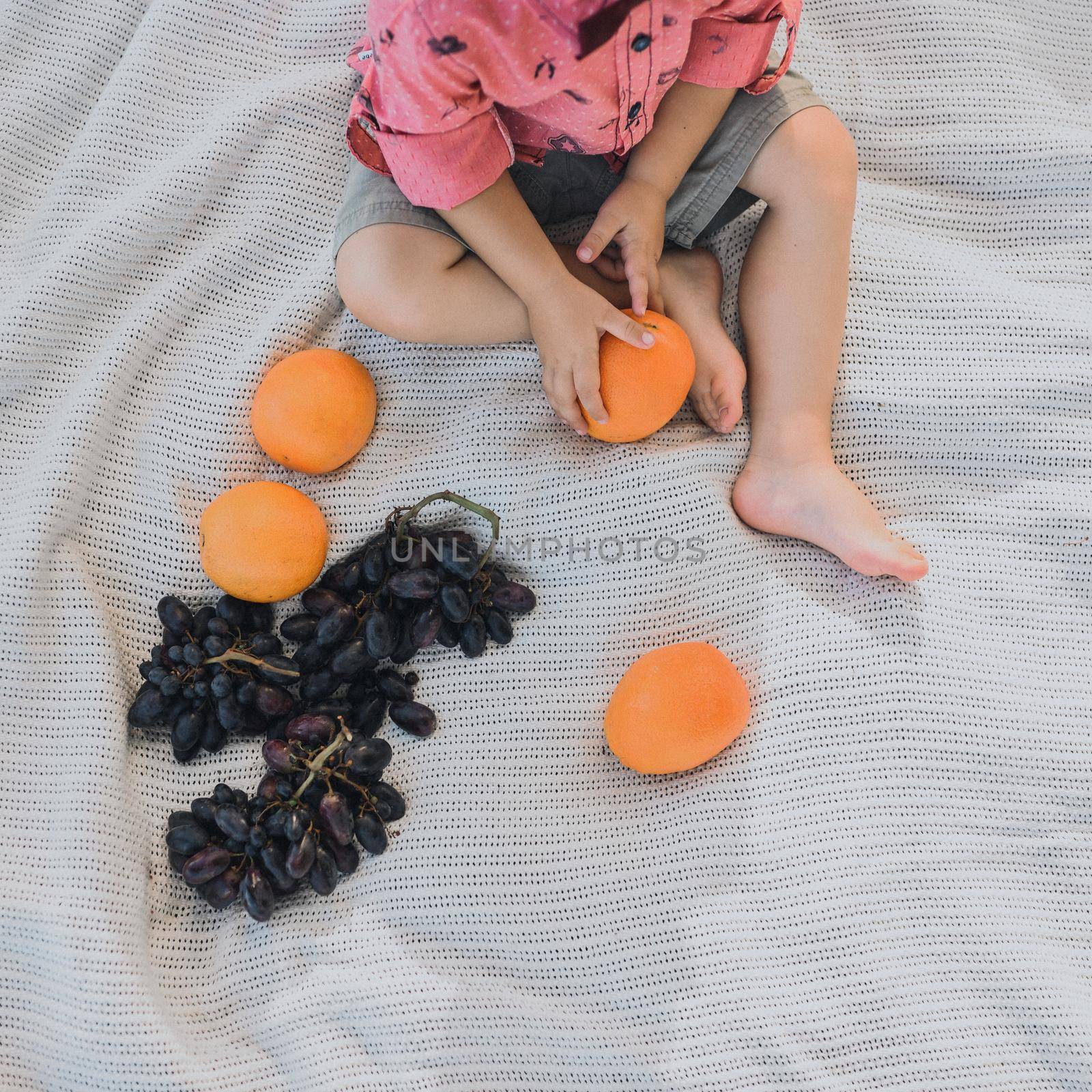 view from above. feet of a boy. holding a bright orange in his hands. Sits on a white bedspread. Picnic in the summer. huge grone of blue grapes.