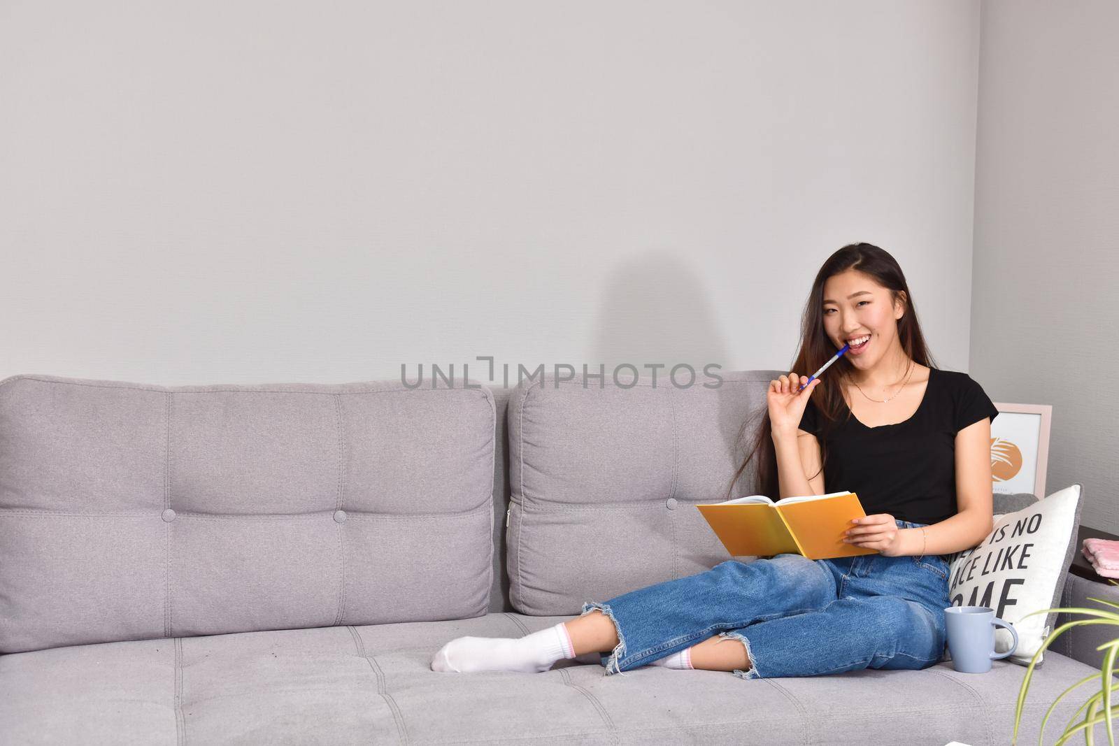 Attractive young asian female with long black hair relaxing on couch at home, writing down ideas for her project, using pen and copybook