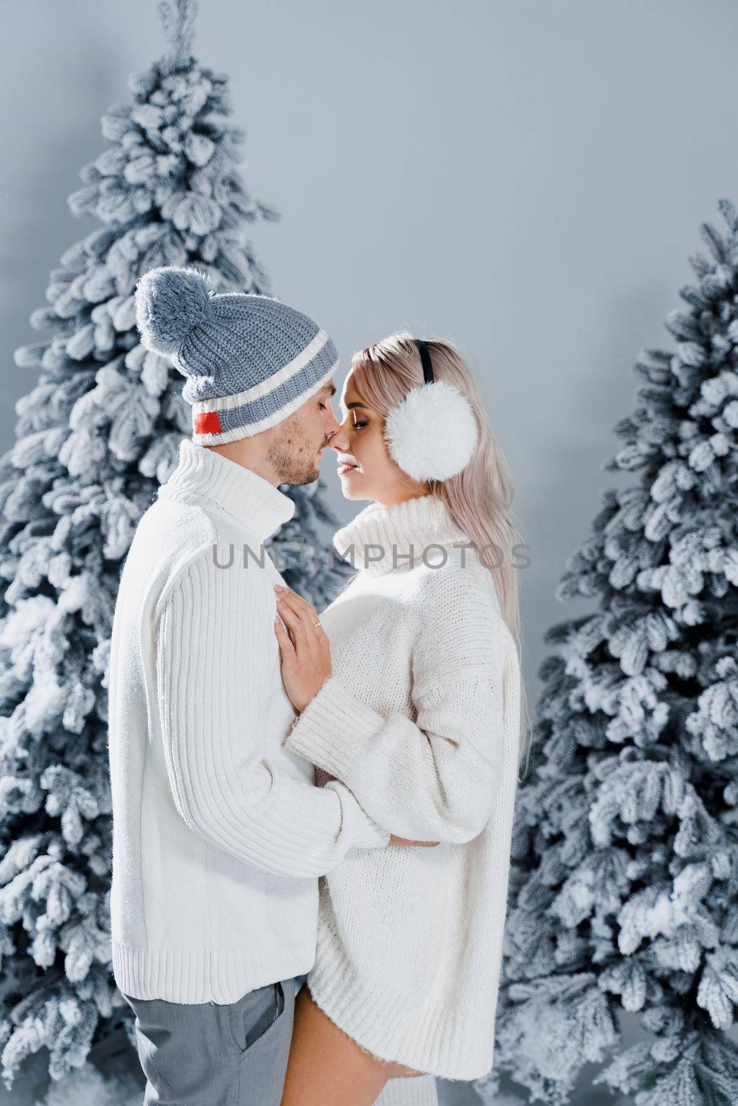 Couple kissing near christmass trees. Winter holidays. Love story of young couple weared white pullovers. Happy man and young woman hug each other by Rabizo