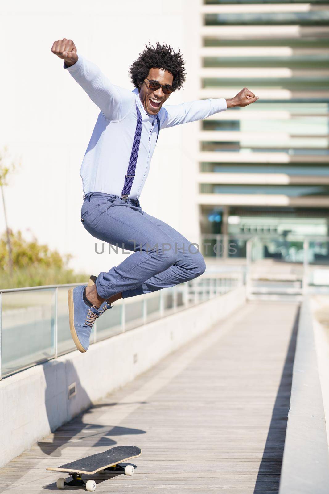 Full body of carefree African American male in smart casual style jumping above skateboard in city and looking at camera