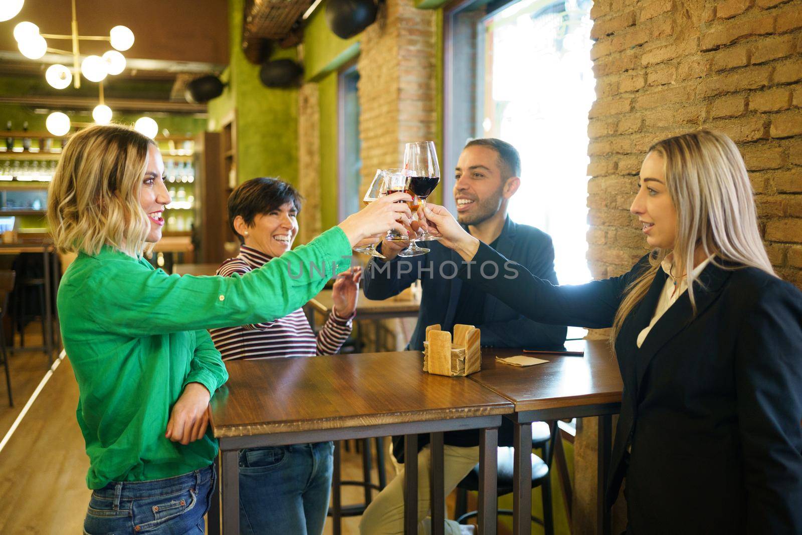 Multiracial man and women smiling and clinking glasses of alcohol beverages over table while proposing toast during meeting in bar