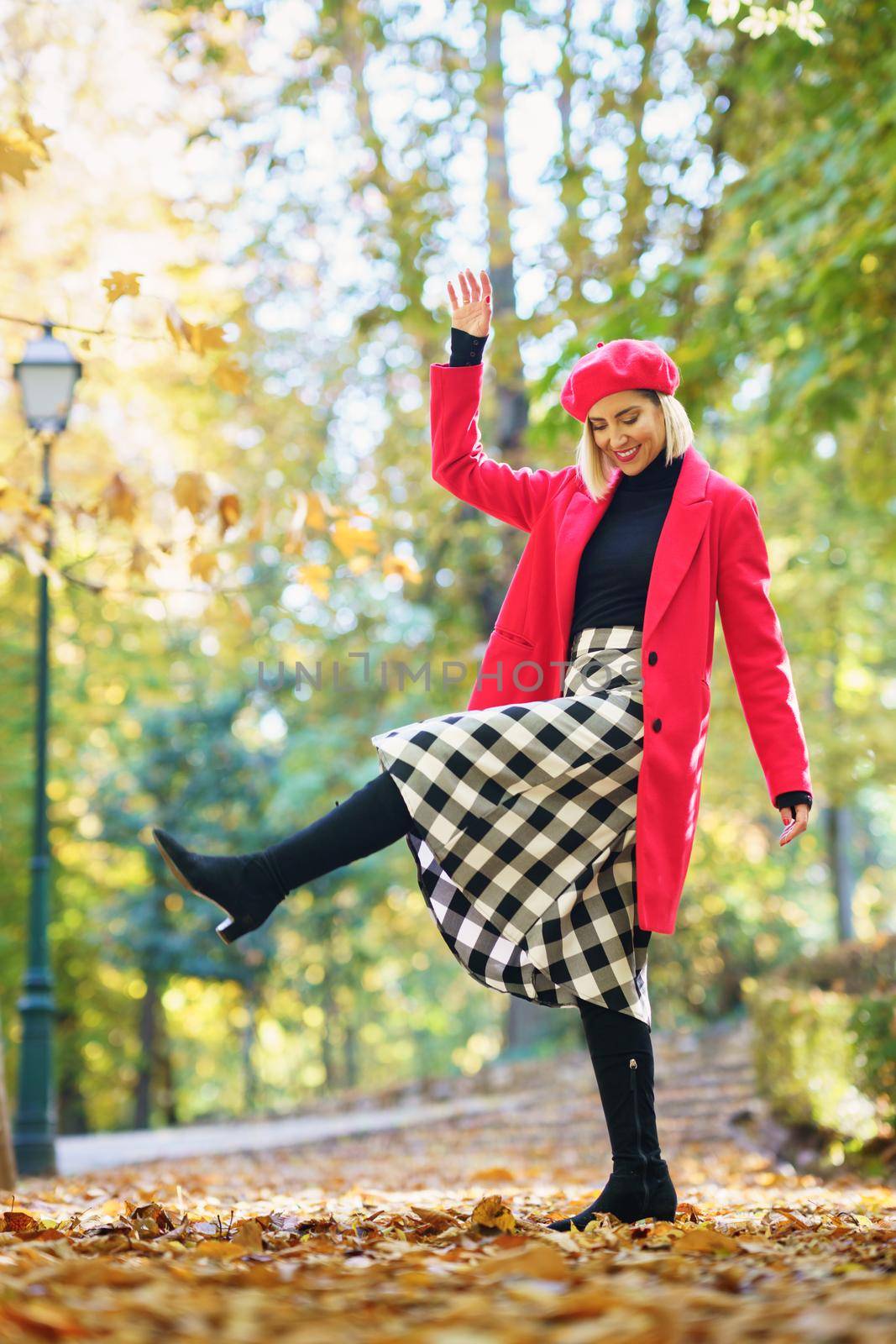 Full body of positive female in red coat and beret raising leg while standing on pathway with fallen leaves in park