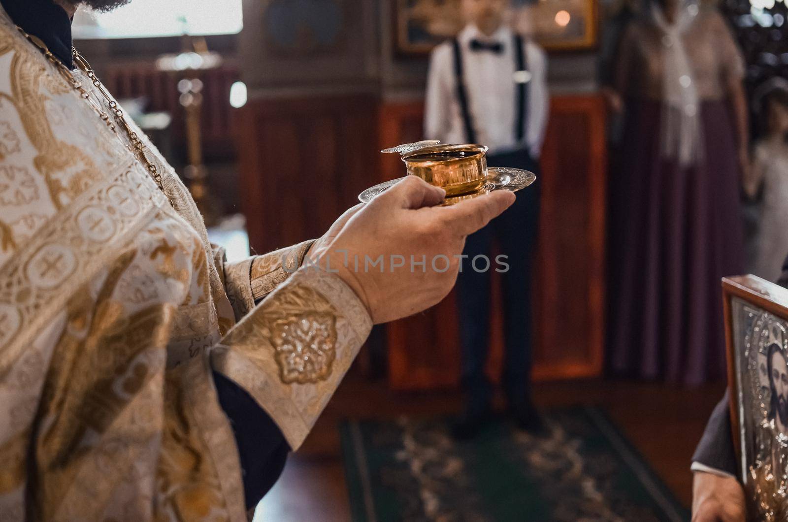 A priest in a golden cassock holds a small mug of sacred red wine on a platter.