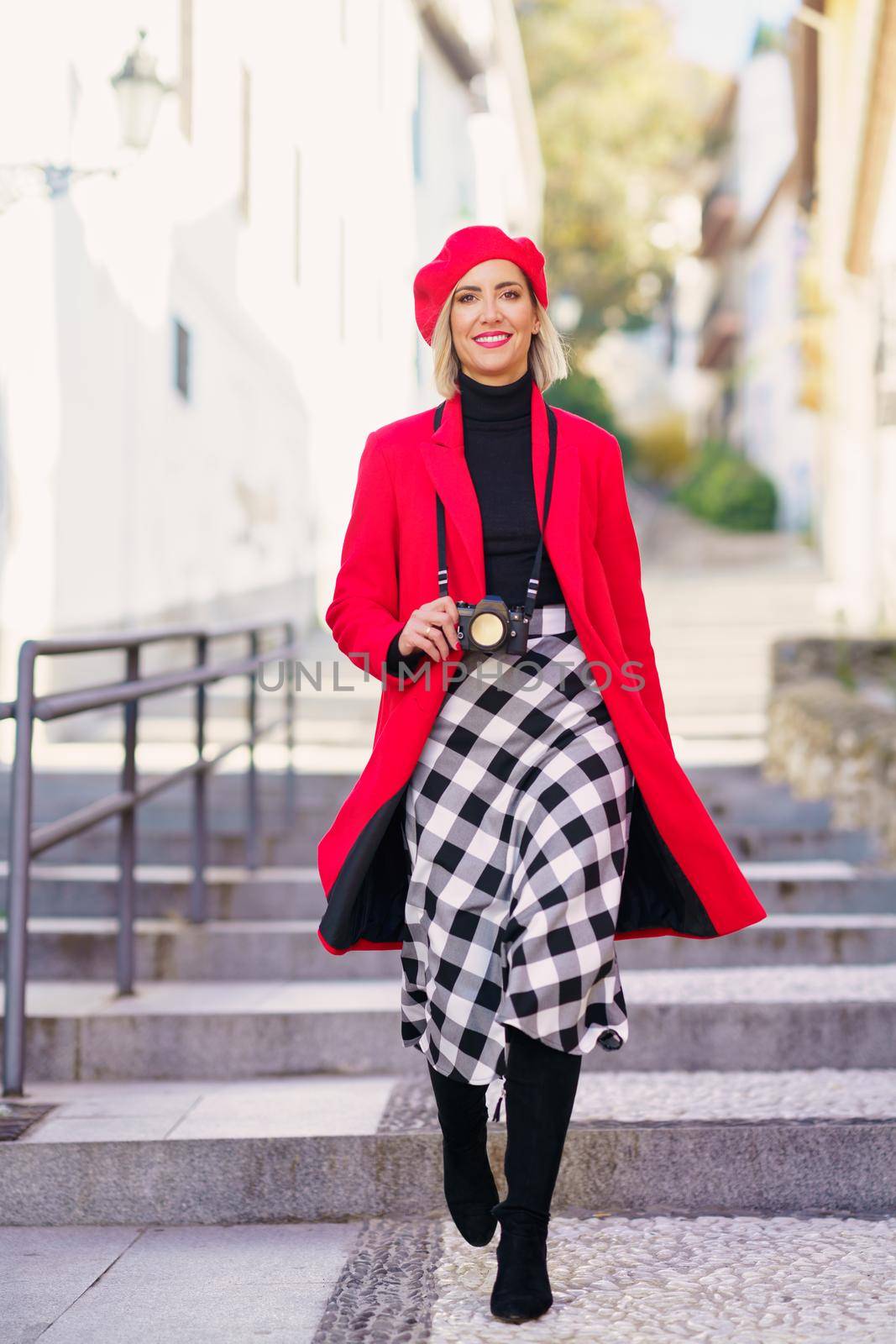 Full length of self assured young lady, with blond hair in elegant outfit and beret, smiling and looking at camera while walking on stone stones with photo camera