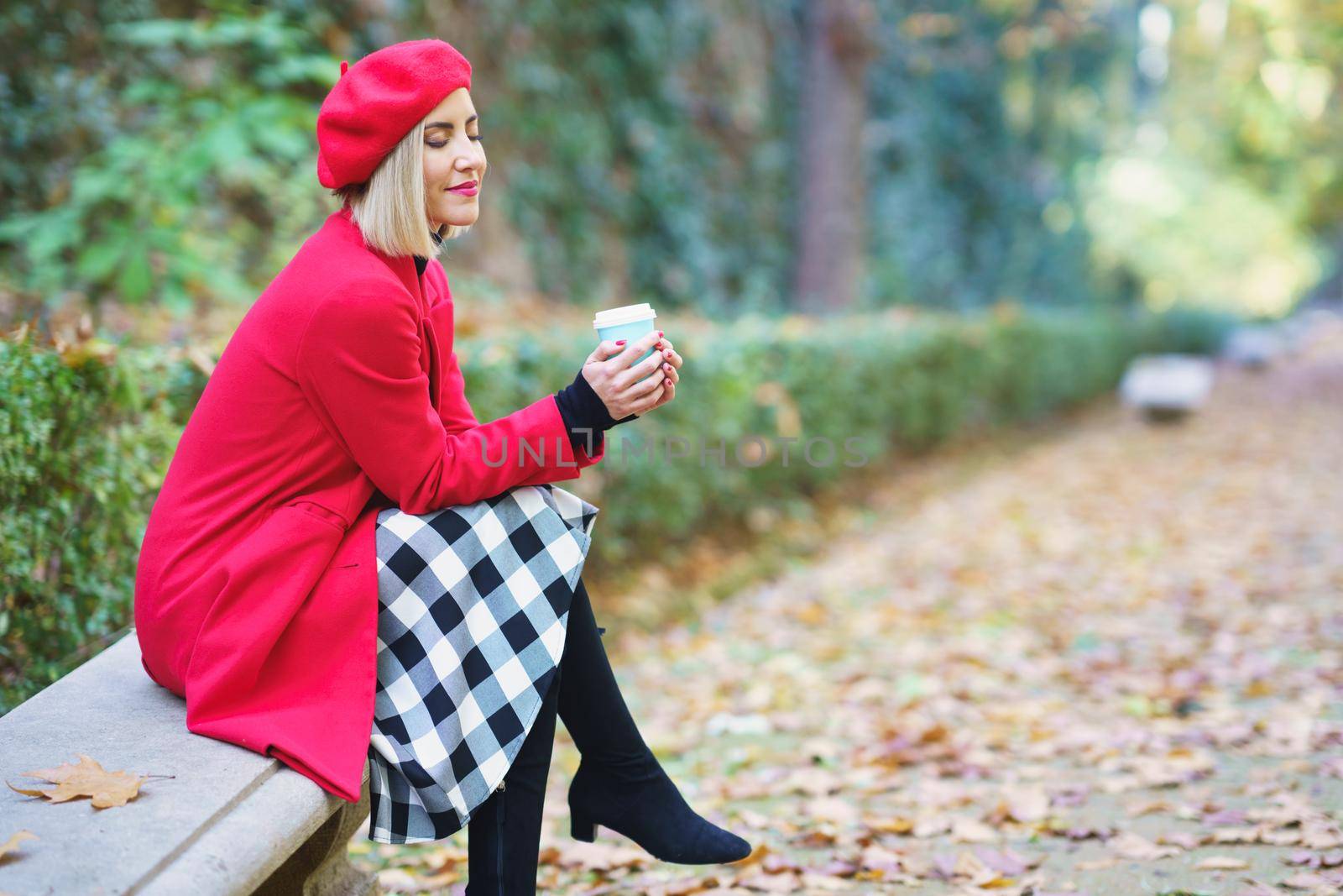 Side view of dreamy female with closed eyes wearing red beret and coat sitting on bench in park with fallen dry leaves and drinking coffee