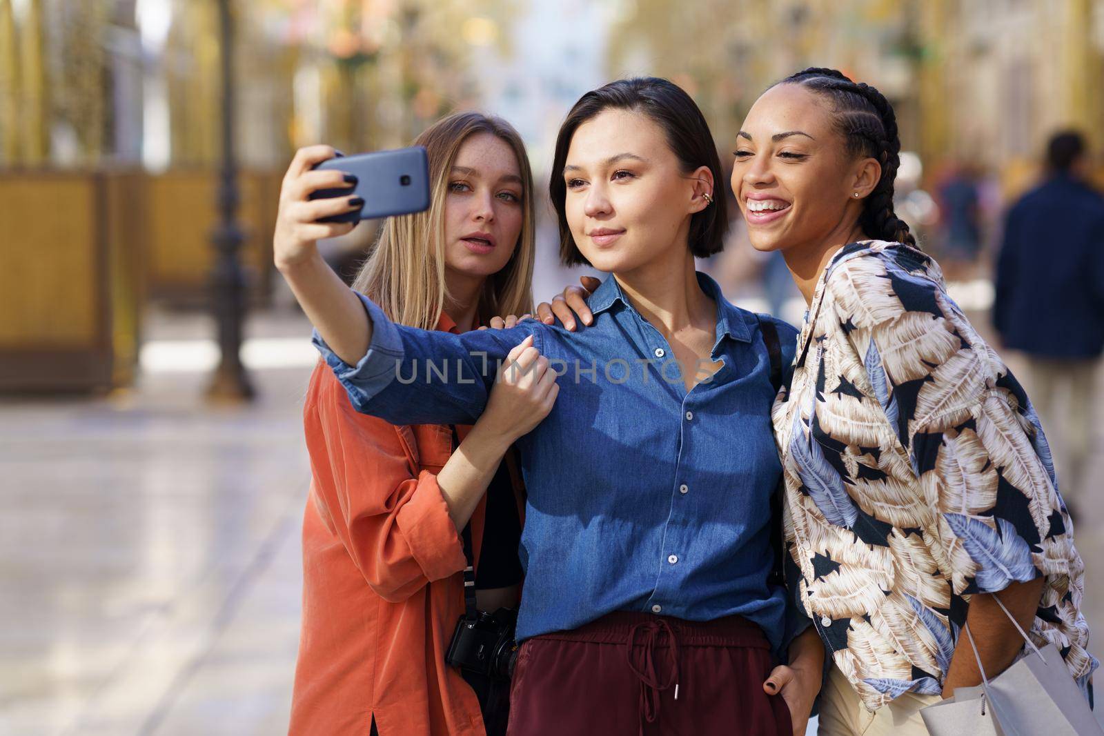 Content multiracial female friends taking self portrait on modern cellphone while standing together on sidewalk against blurred background of city