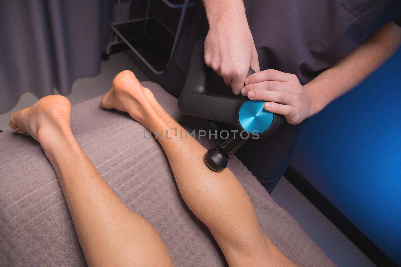 A professional masseur makes a massage of the calf muscle with a massage gun to a man athlete in a massage room with low light by yanik88