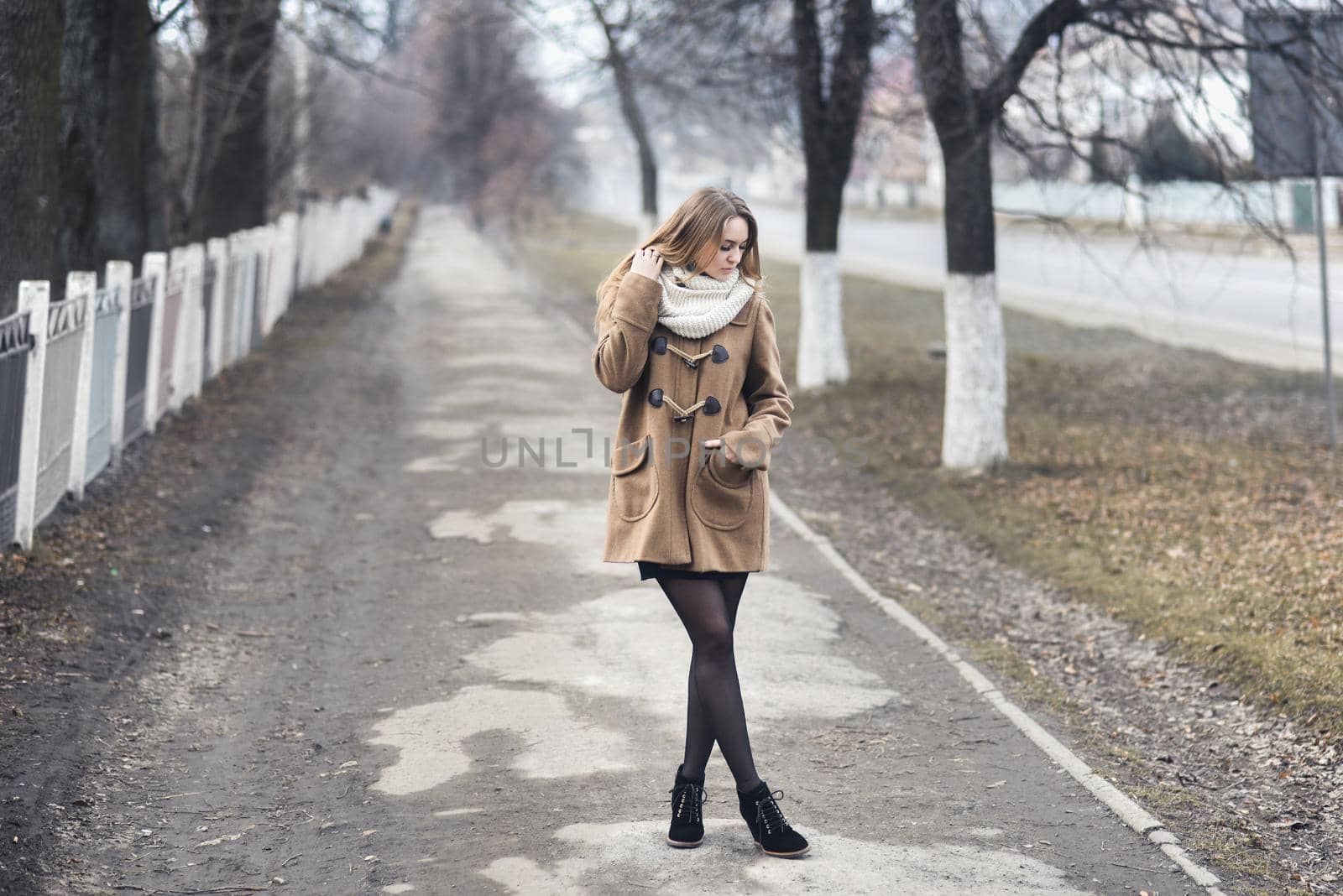 Blond-haired young girl cheerful joyful dressed you in a beige coat and white scarf. Autumn gray street on the background. empty road