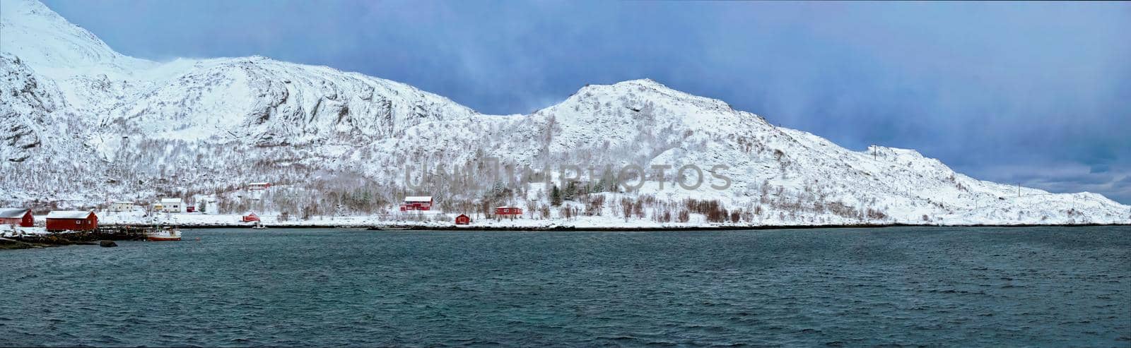 Panorama of Norwegian fjord with traditional red rorbu houses on fjord shore in snow in winter. Lofoten islands, Norway