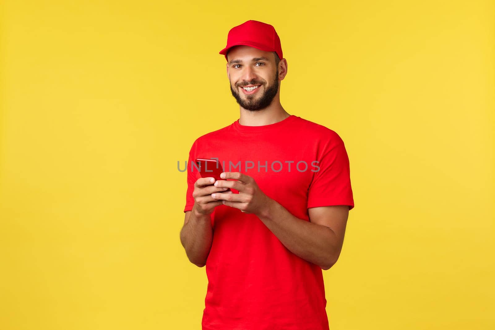 Contactless payment, online delivery, internet shopping and parcel tracking concept. Friendly smiling courier in red uniform cap, t-shirt, using mobile phone and look camera, yellow background.