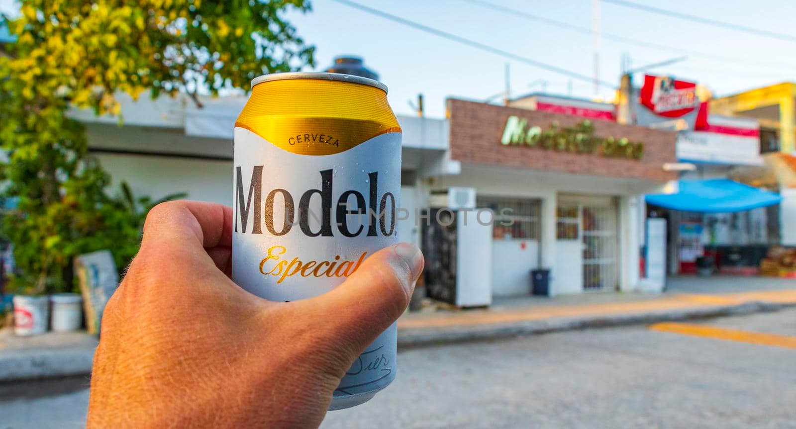Chemuyil Mexico 02. February 2022 Modelo beer can in the hand with cityscape landscape and store of Chemuyil in Quintana Roo Mexico.
