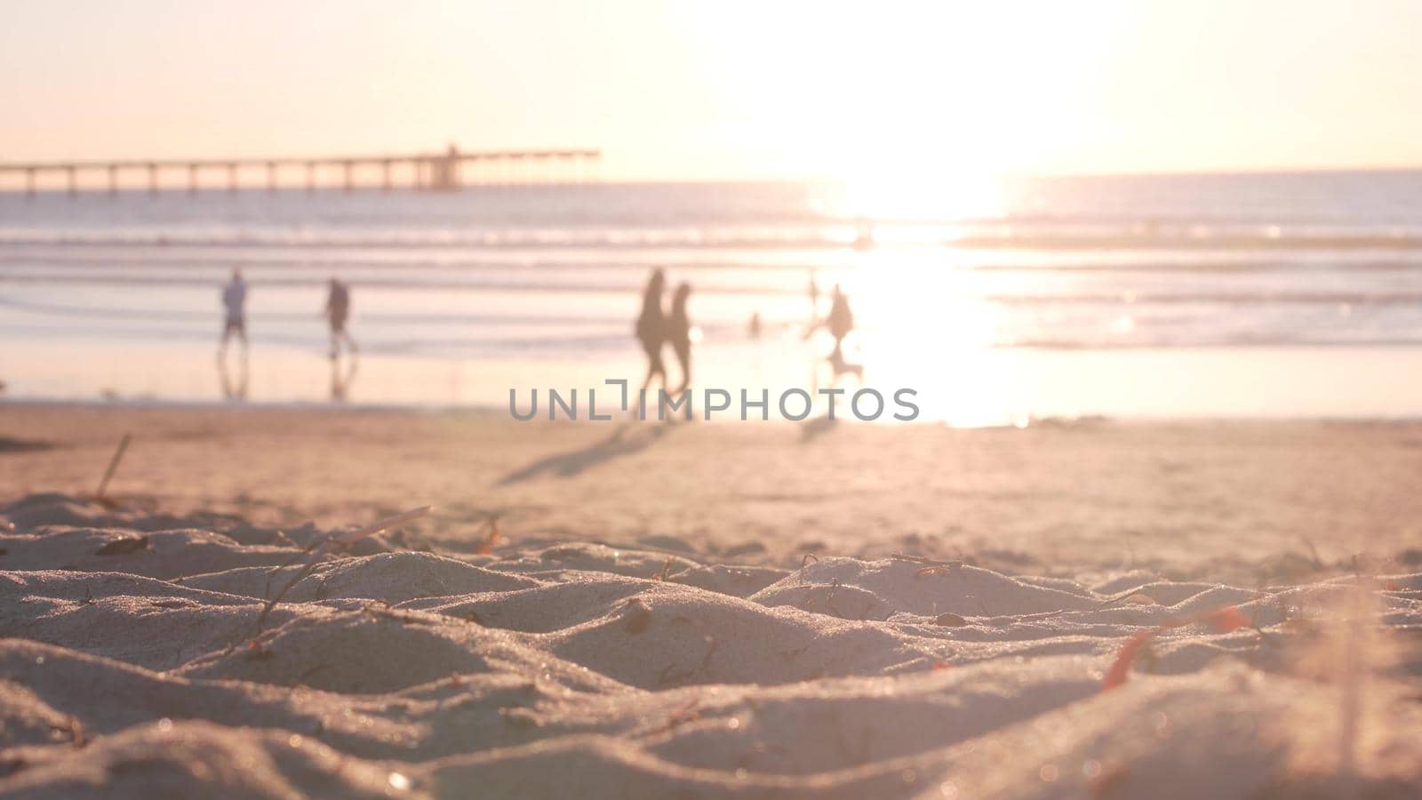 People walking on sandy Ocean Beach by pier at sunset, California coast, USA. Unrecognizable defocused family silhouettes in bright sun light, vacations on shorein sunshine. Sunlit sea water waves.