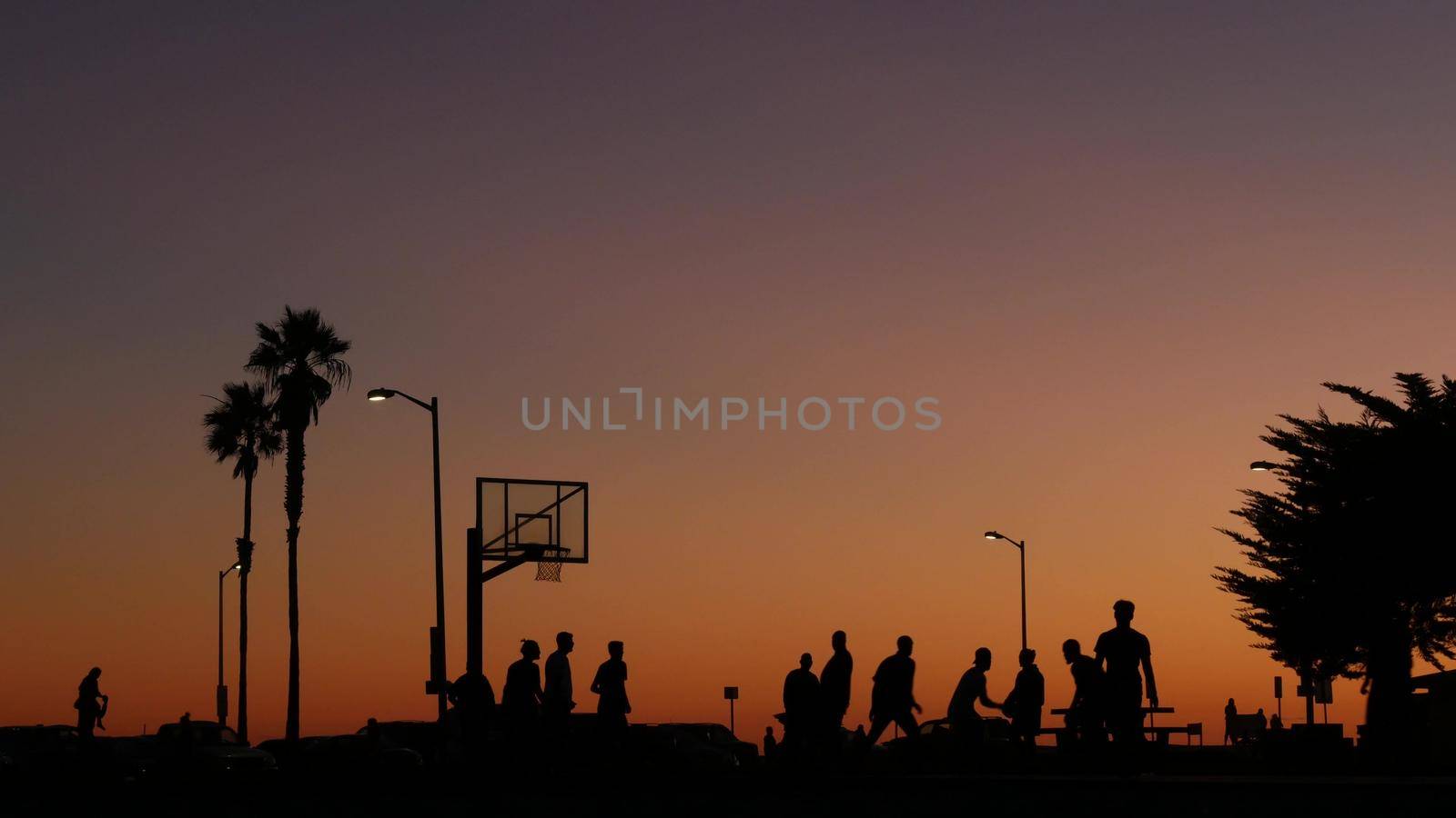 People on basketball court playing basket ball game. Sunset on beach, California by DogoraSun