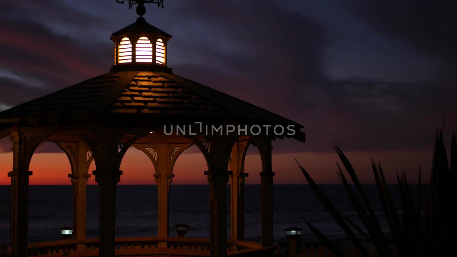 Sunset cloudscape and gazebo silhouette on beach, twilight dusk, clouds in dramatic purple pink sky, California coast. Wooden romantic cosy alcove and palm tree. Weather wind vane or roof weathercock.
