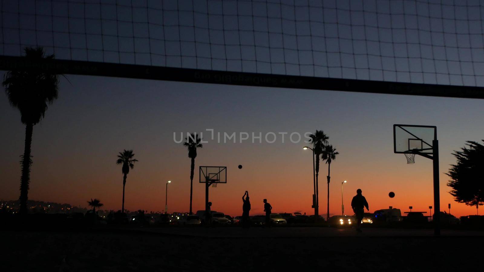 Silhouettes of players on basketball court outdoor, people playing basket ball game, sunset ocean beach, California coast, USA. Black hoop, net and backboard on streetball sport field. Mission beach.