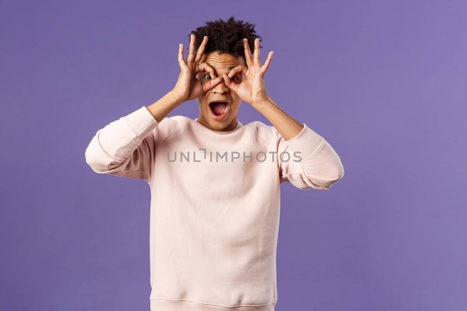 Portrait of funny, enthusiastic young hispanic man fooling around, making finger glasses, mask with hands over eyes, open mouth fascinated and surprised, seeing great product or promo action.