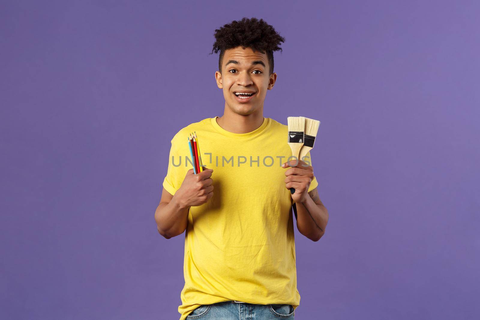 Portrait of upbeat young creative man, artist starting art courses, want to learn how to draw, holding colored pencils and brushes, standing enthusiastic over purple background.