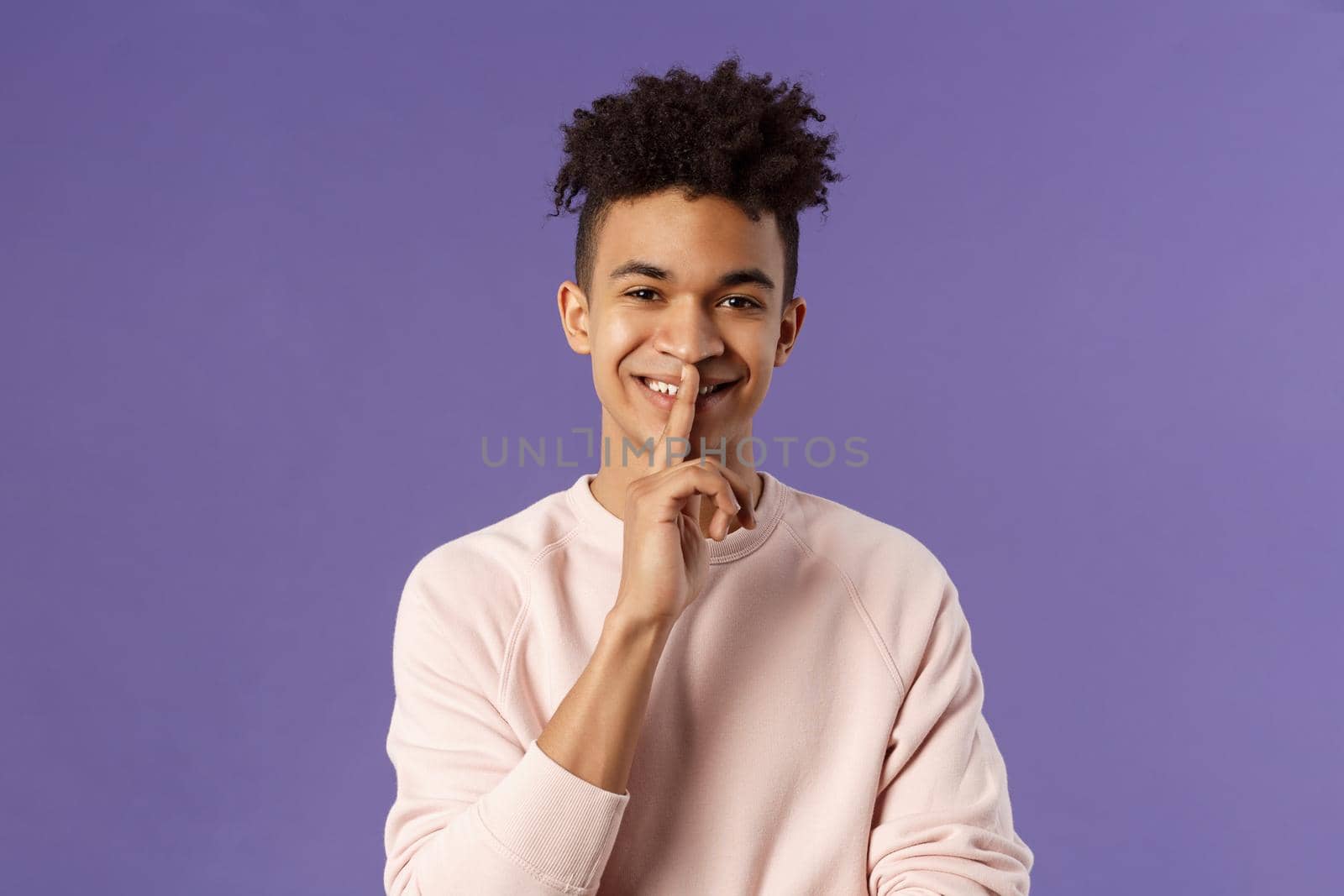 Close-up portrait of attractive smiling young man hiding secret, asking keep silent or quiet to do surprise, show shush gesture, place index finger to mouth, standing purple background.