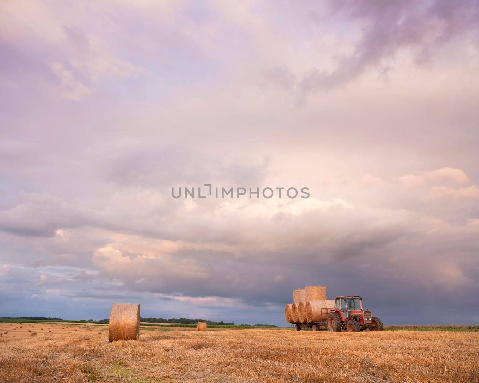 landscape with straw bales and tractor in french ardennes under cloudy sky during sunset in summer